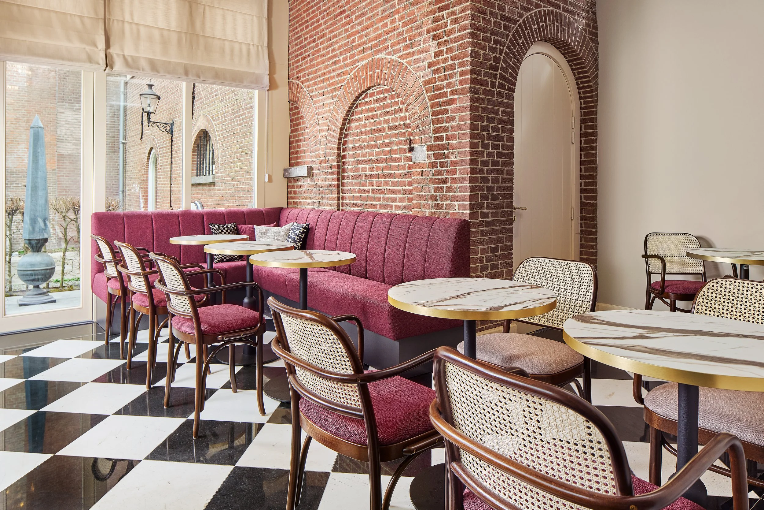 Interior of a modern café with round marble tables, chairs with wooden frames and dark velour upholstery, a red sofa along large windows overlooking an outdoor area with antique ornaments, and exposed brick walls.