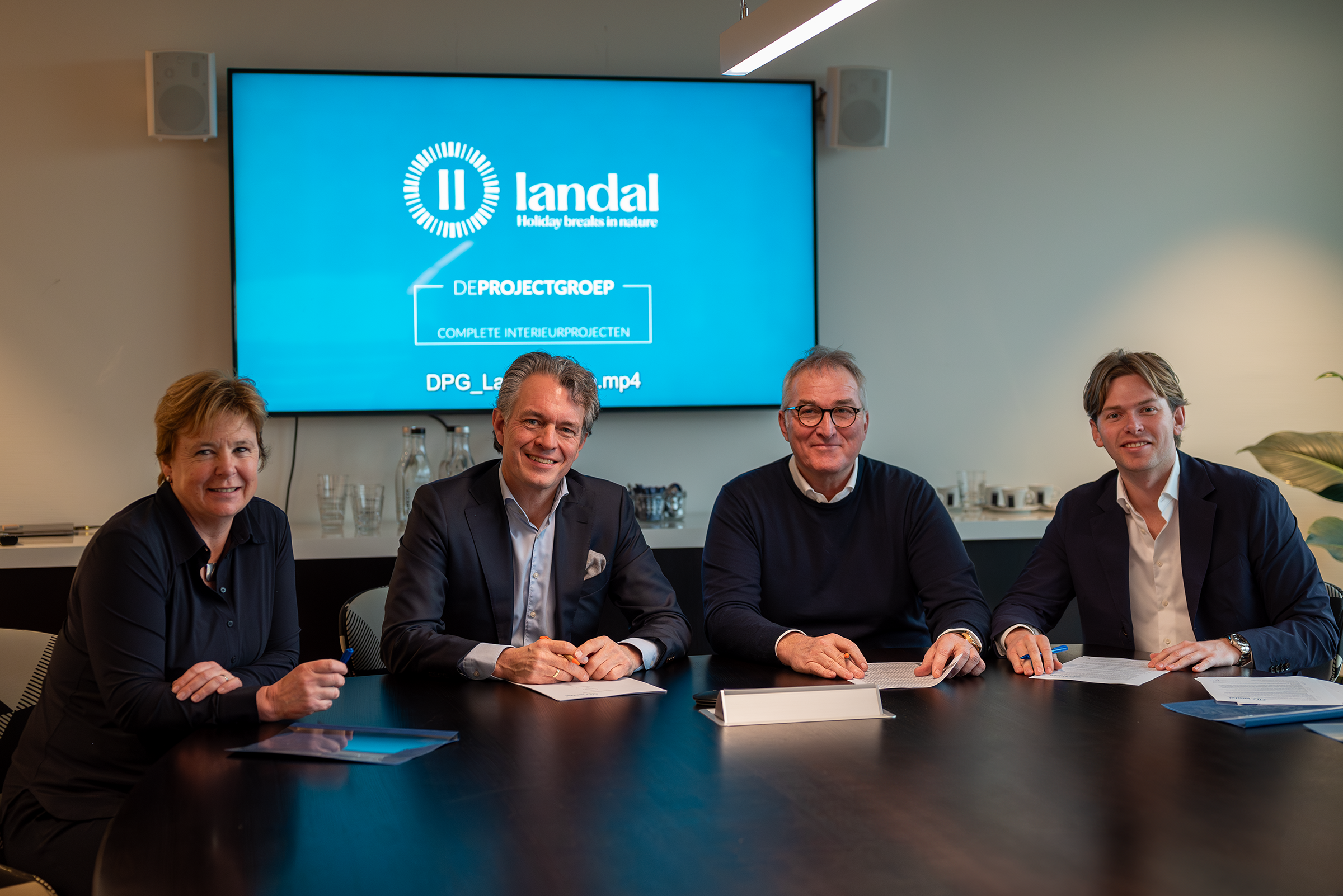 Vier mensen zitten naast elkaar aan een tafel tijdens een zakelijke bijeenkomst, met een grote scherm op de achtergrond dat het logo van Landal en zakelijke tekst toont.