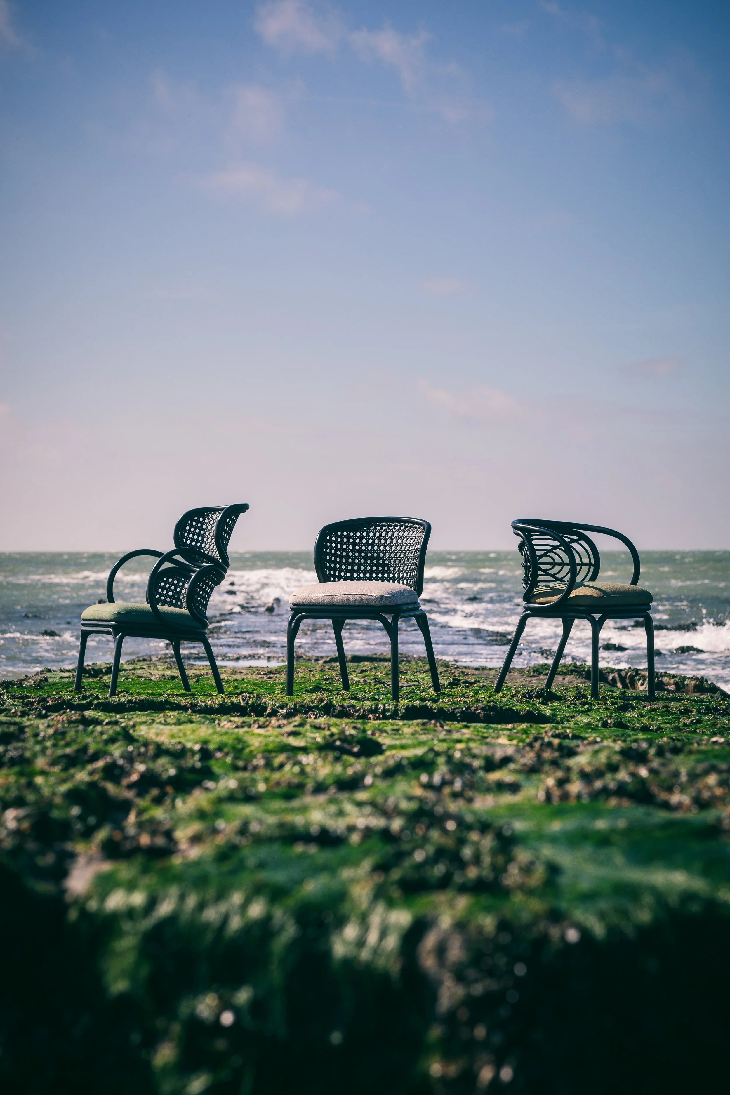 Three black wicker chairs with cushions on a grassy surface in front of the ocean, under a clear blue sky with a few clouds.