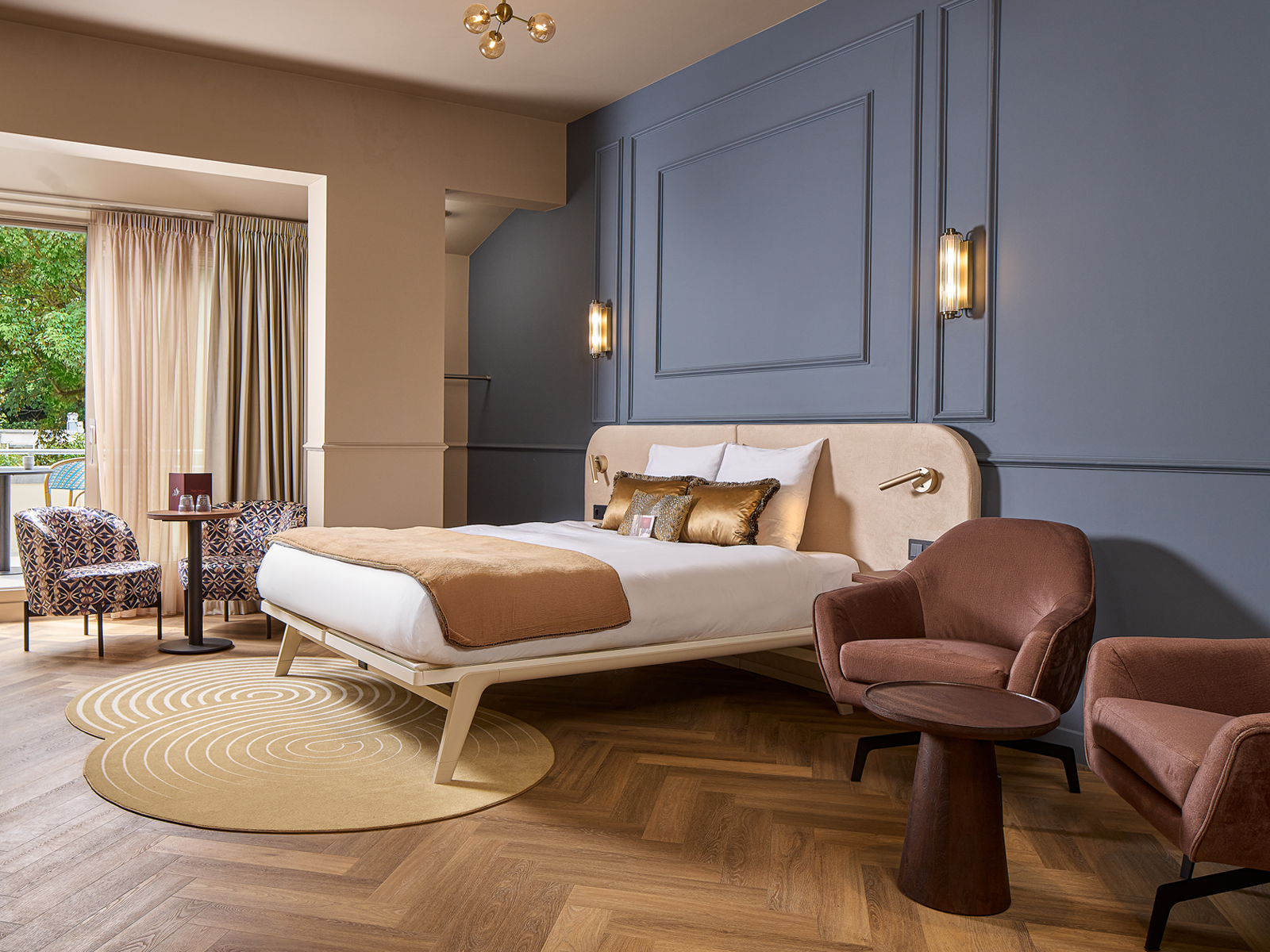 A hotel room with a large bed, two armchairs, a small table, and a balcony overlooking green trees.