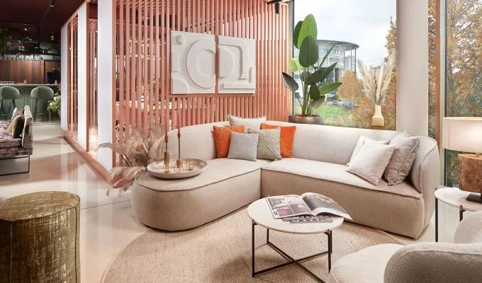 Bright living room with a large L-shaped sofa, decorative cushions, a round coffee table with an open book, large green plants, and large windows with a view outside.