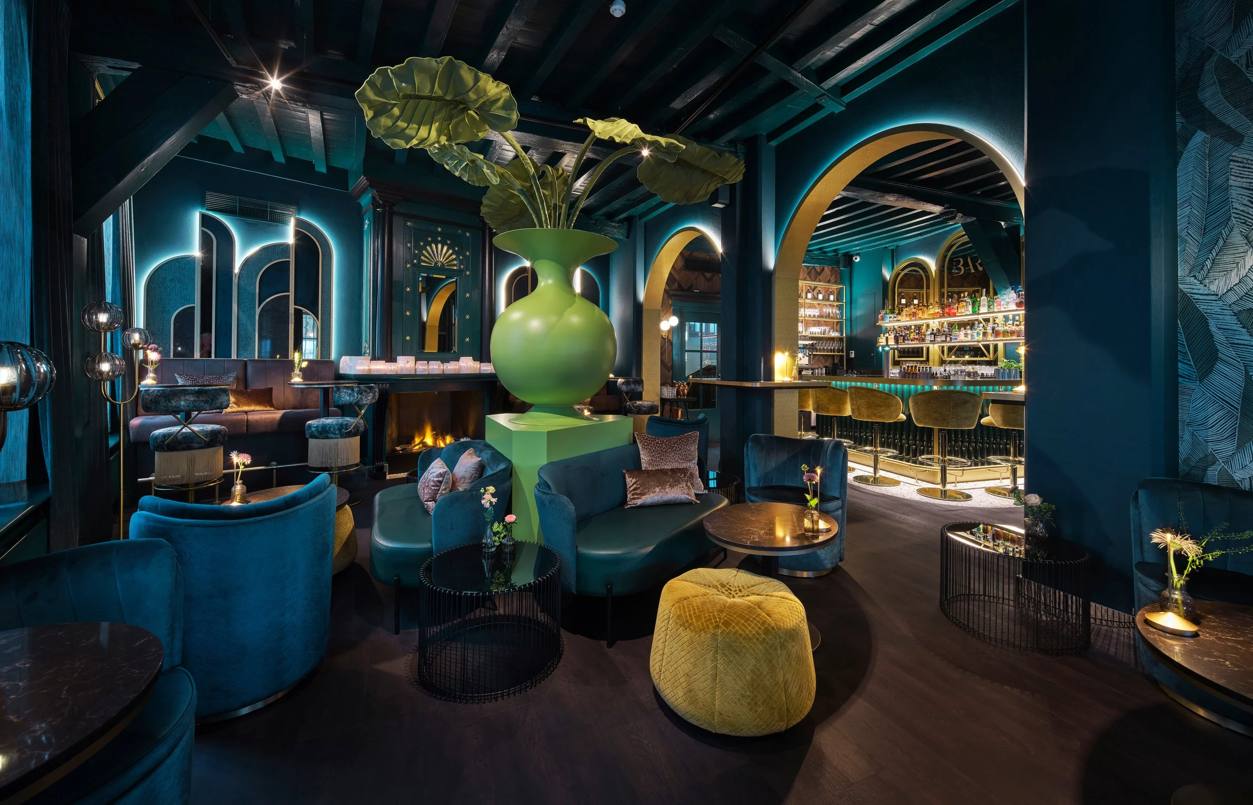 A luxurious, modern bar and lounge area with blue and green furniture, a large green plant in a vase, and an illuminated bar in the background.