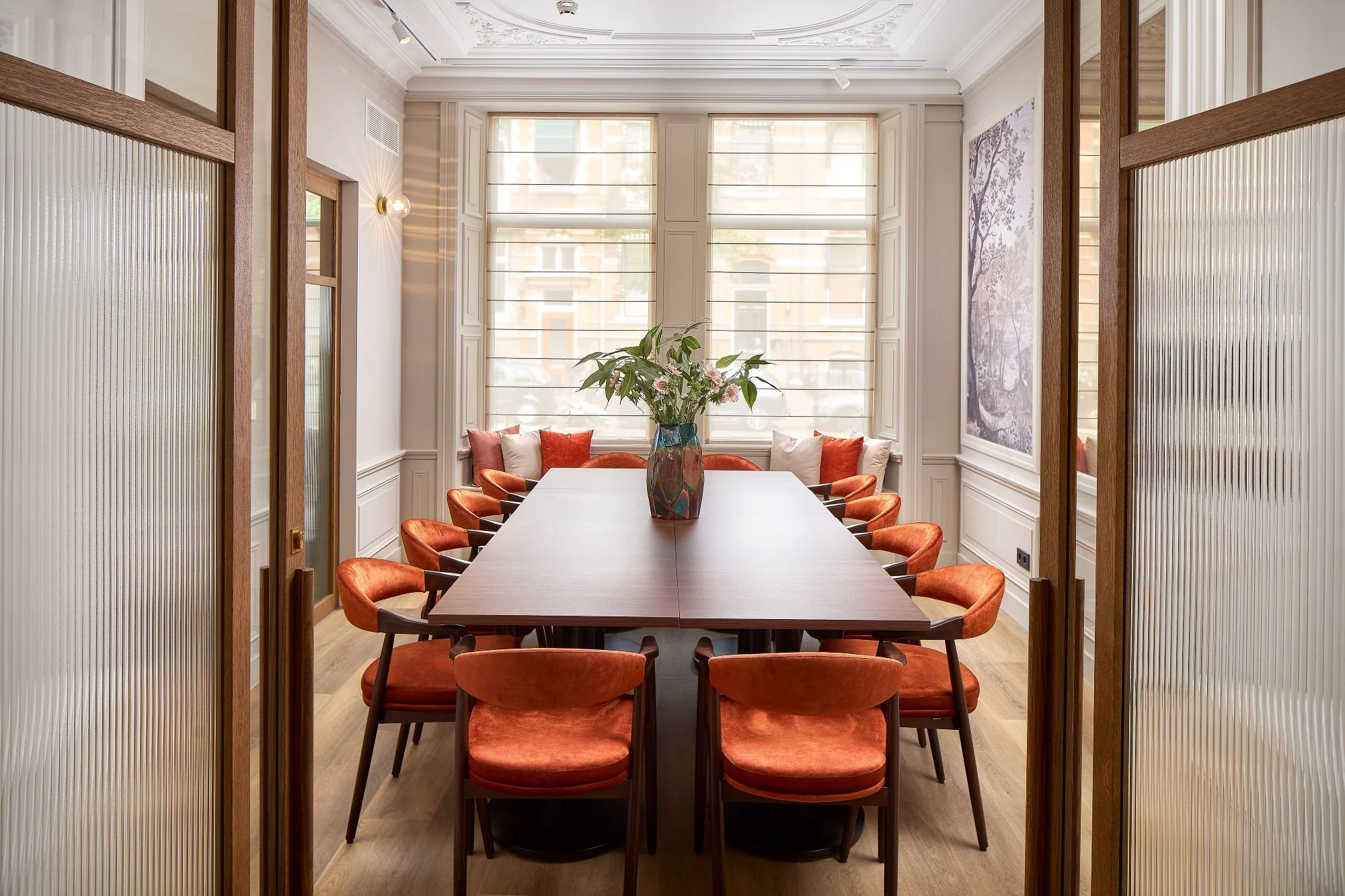 A meeting room with a large table, pink-orange chairs, a large vase with flowers, large windows, and light-colored walls.