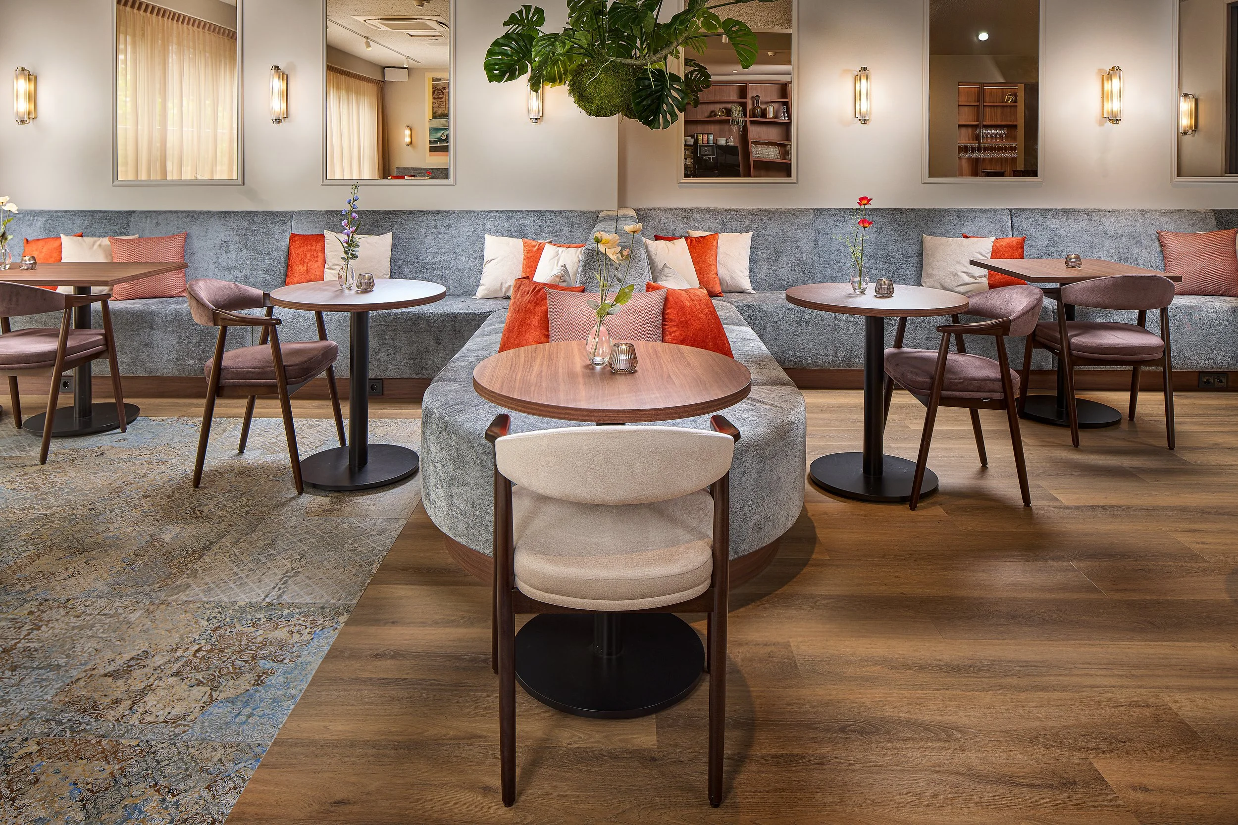 Interior of a chic restaurant or café with round wooden tables, comfortable chairs, and a large flowering plant. The wall is decorated with mirrors and atmospheric wall lighting.
