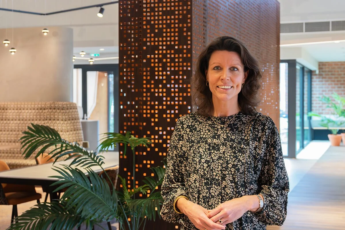 Vrouw met donker gekleurde, bloemenpatroon blouse in een moderne, sfeervolle binnenruimte met planten, grote ramen en decoratieve muurpanelen.