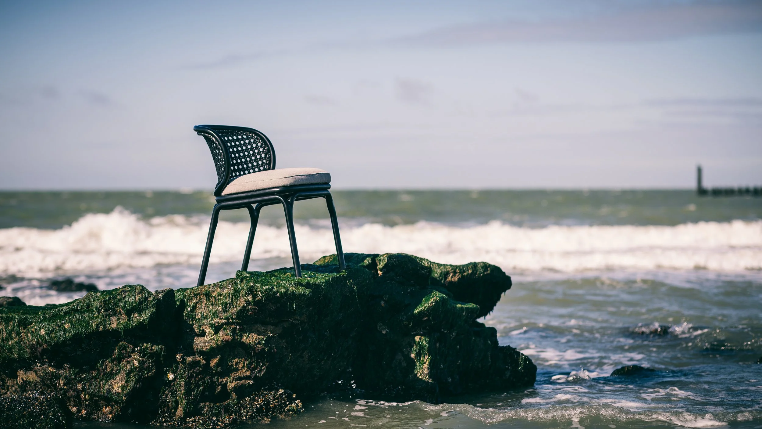 Een stoel staat op rotsen aan de kust bij de zee, met golven en een blauwe lucht met wolken op de achtergrond.