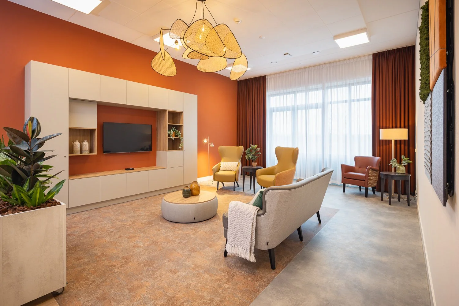 Modern living room with orange-red accent wall, comfortable chairs, a television, large windows with white and orange curtains, and decorative plants.