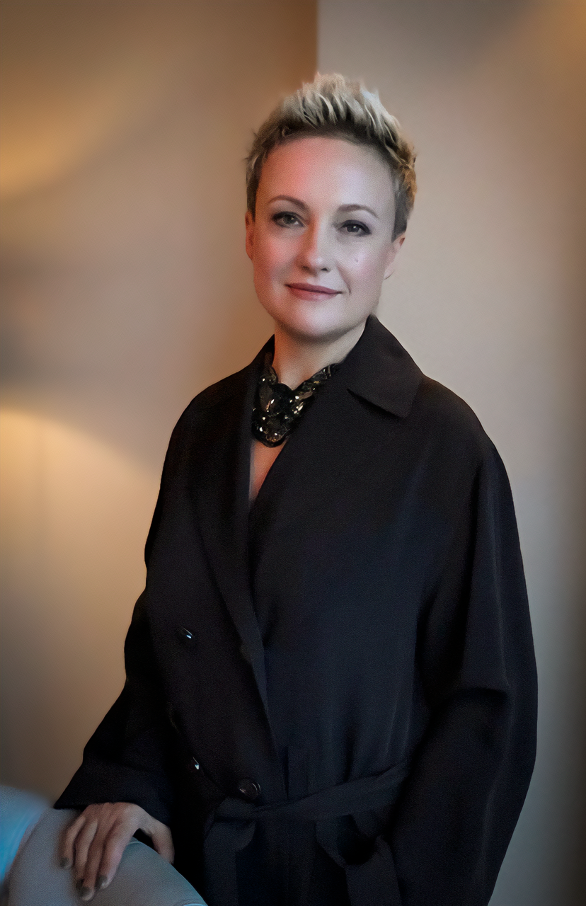 A woman with short, blonde hair styled upward, wearing a black trench coat and a decorative black necklace, standing indoors against a neutral background.