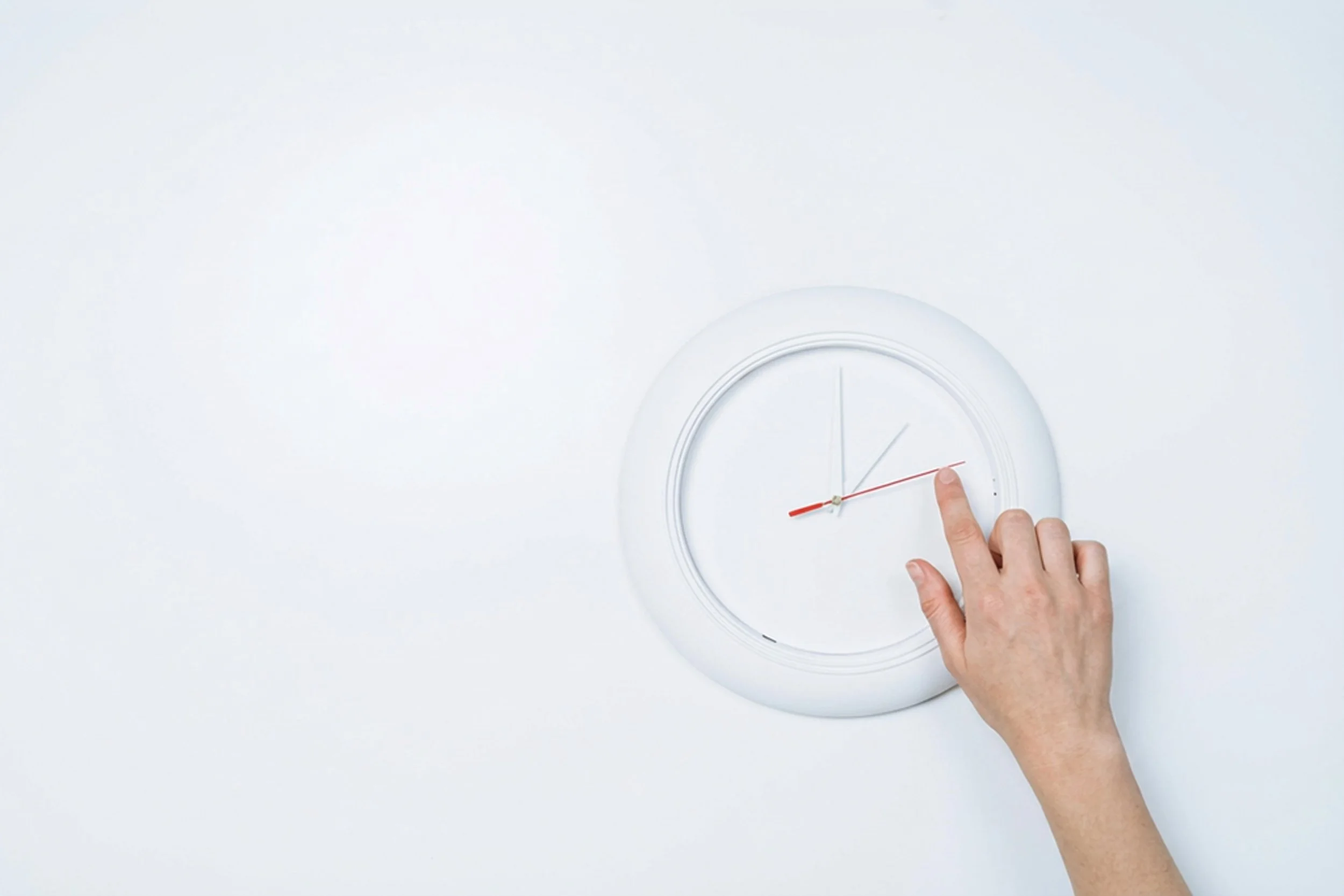 A white wall clock with black hour and minute hands and a red second hand is mounted on a white wall. A person's hand is touching the clock near the 3 o'clock position.