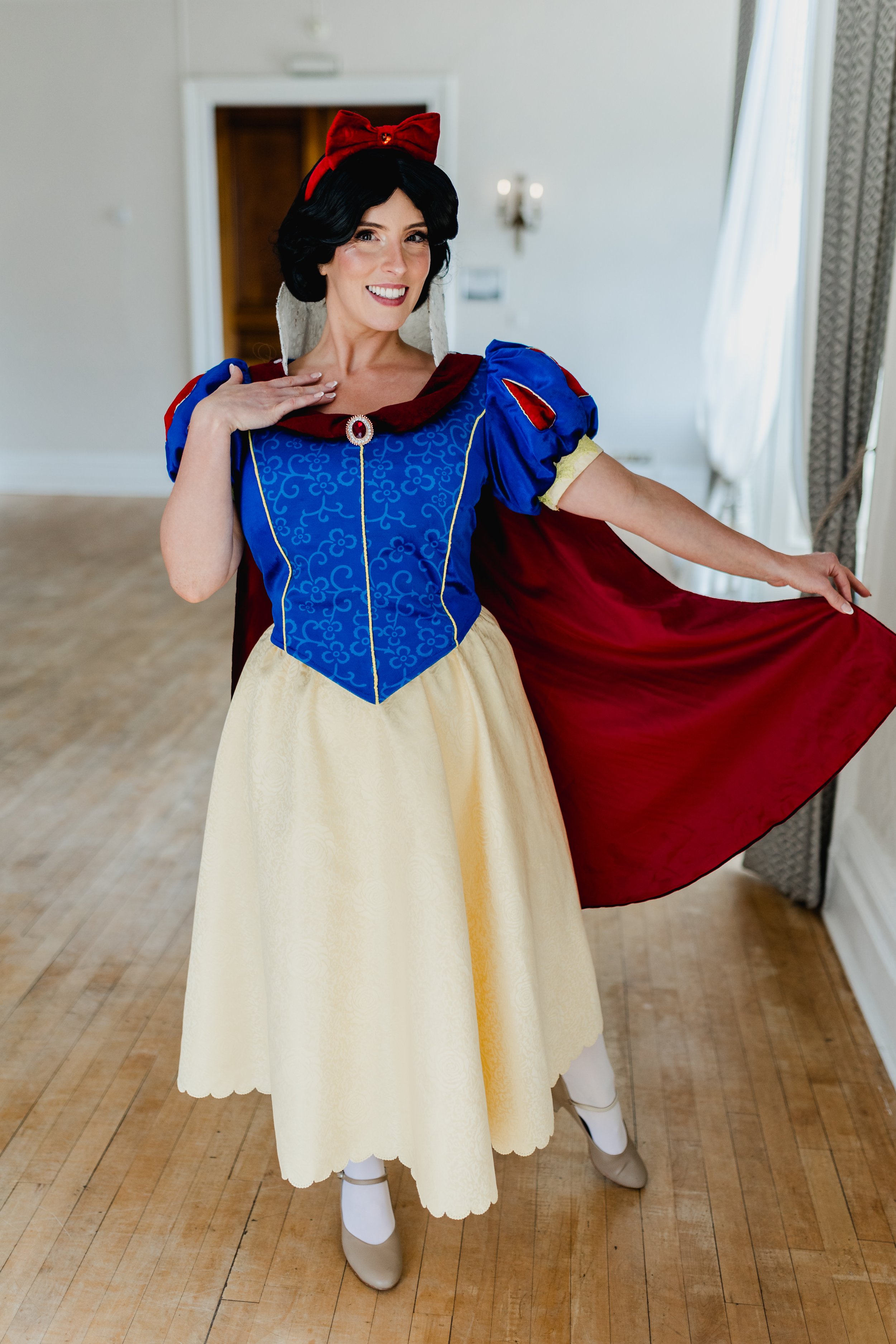 Woman in Snow White costume with blue bodice, yellow skirt, red cape, and red bow headband, smiling in room with wooden floor and curtains.