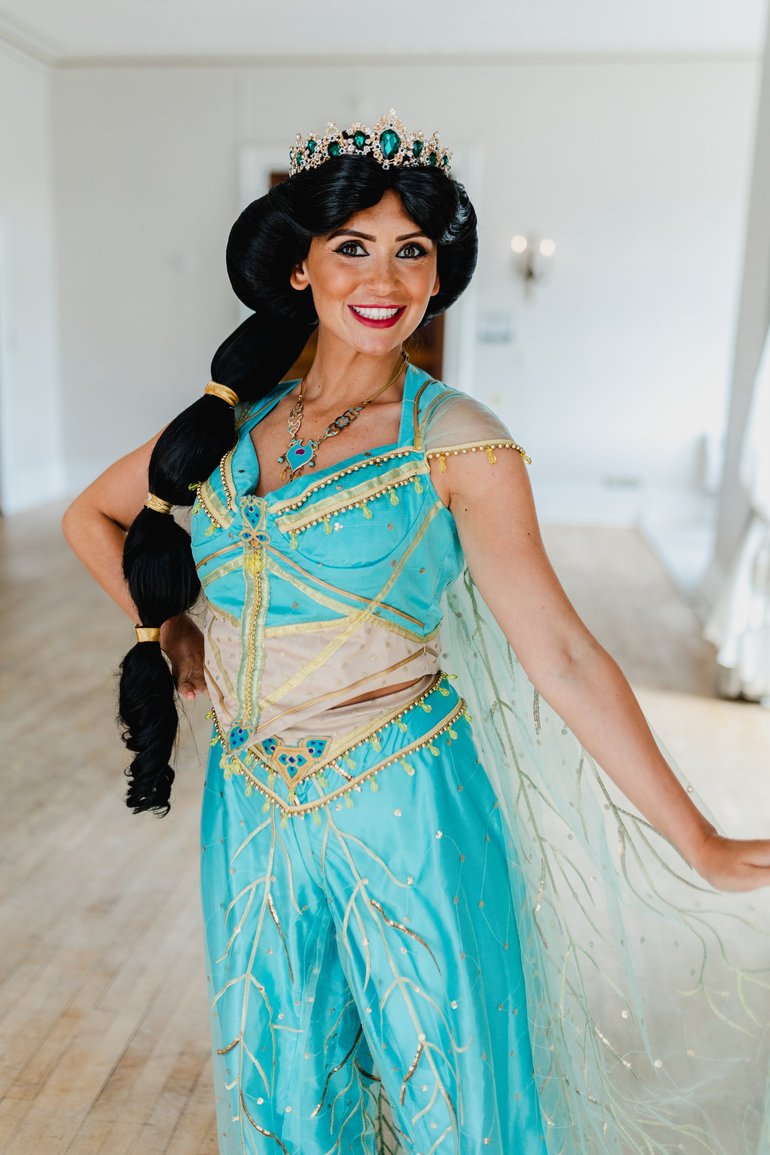 A woman dressed as Princess Jasmine from Aladdin, wearing a blue and gold costume with a tiara, in a bright room.