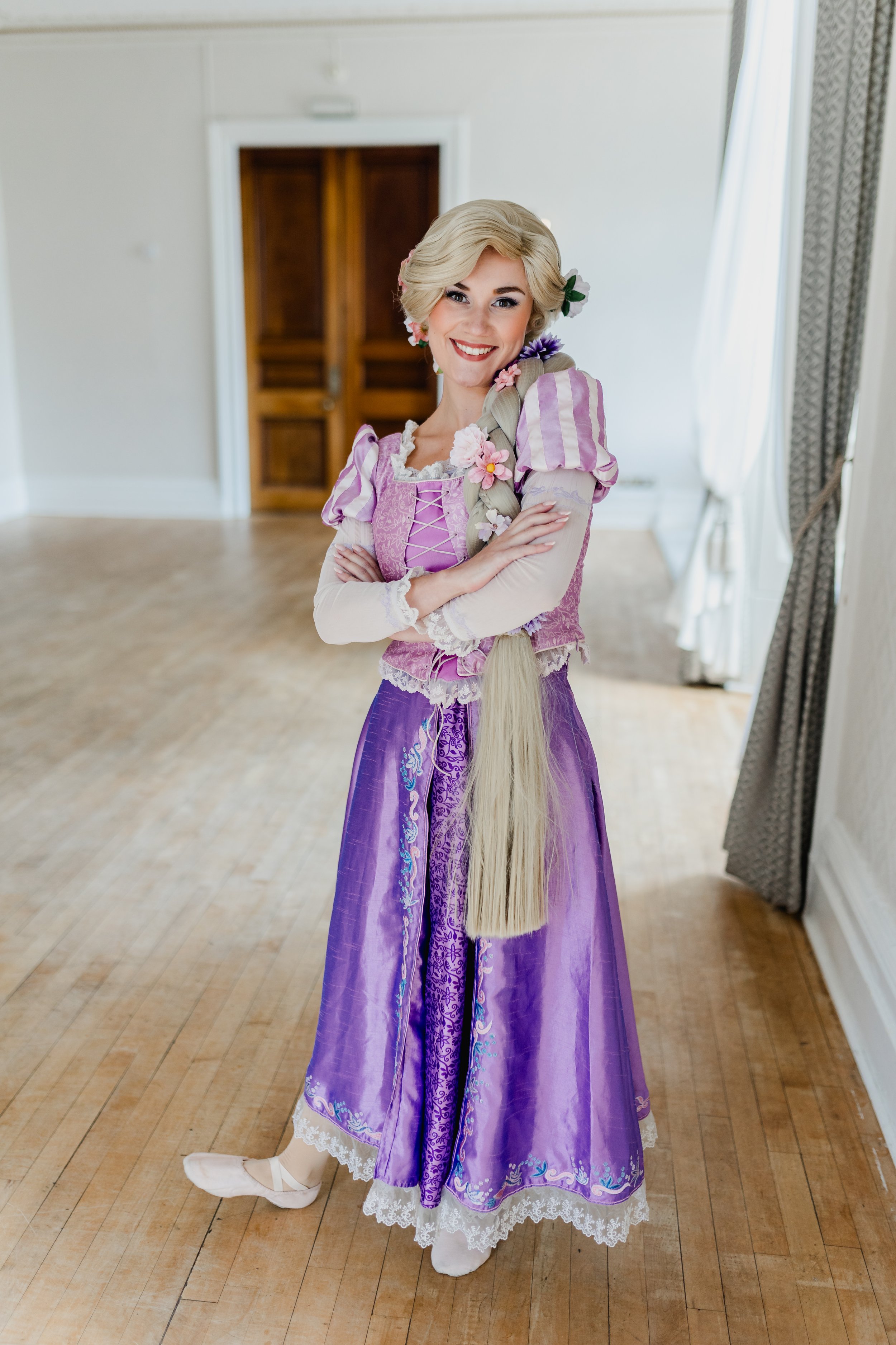 A woman dressed as Rapunzel, wearing a purple and pink medieval-style gown with long blonde hair styled in a braid with flowers, standing with arms crossed and smiling in a room with wooden flooring and white walls.