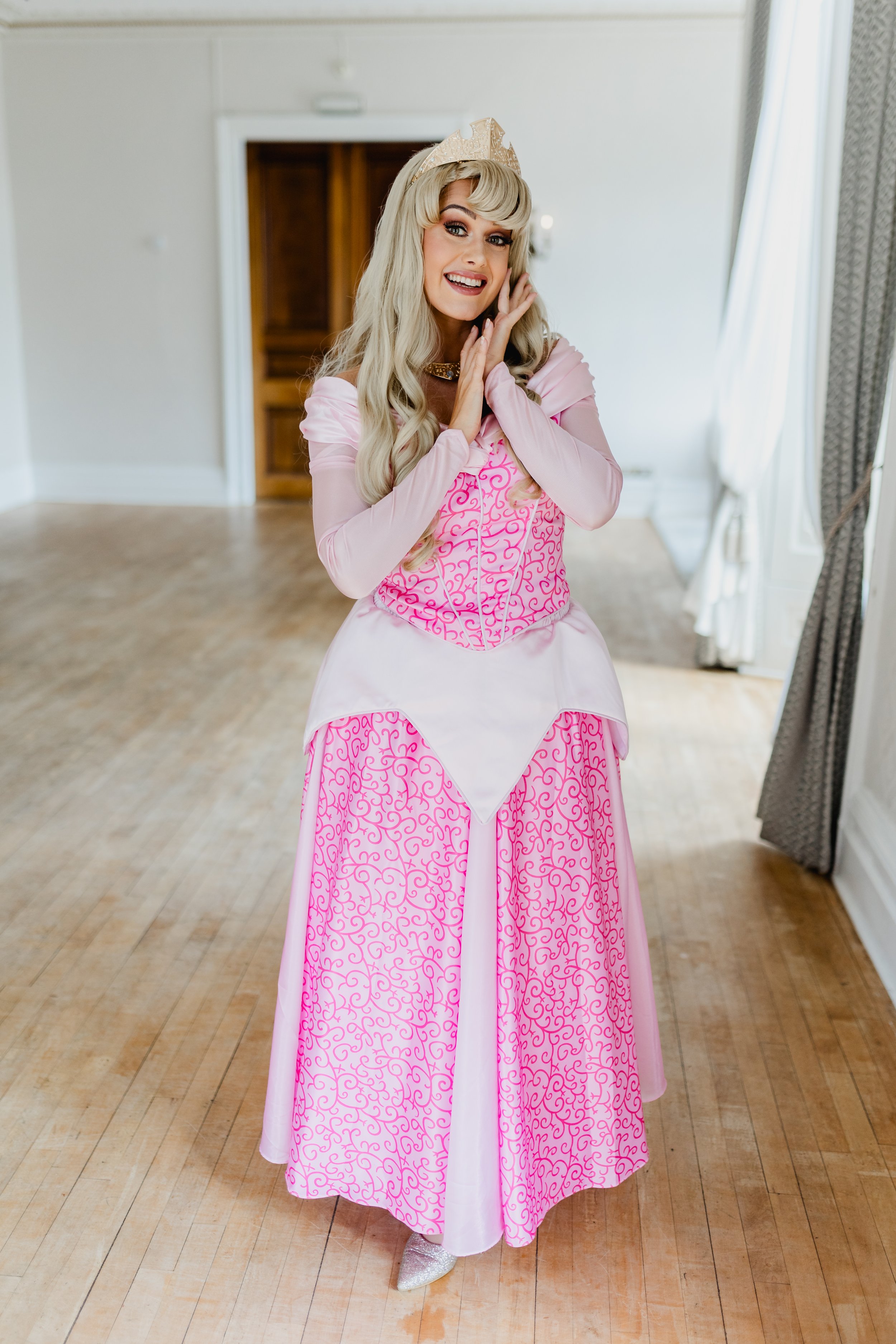 Woman dressed as a princess in a pink and purple gown, wearing a crown, in a room with wooden floors and white walls.