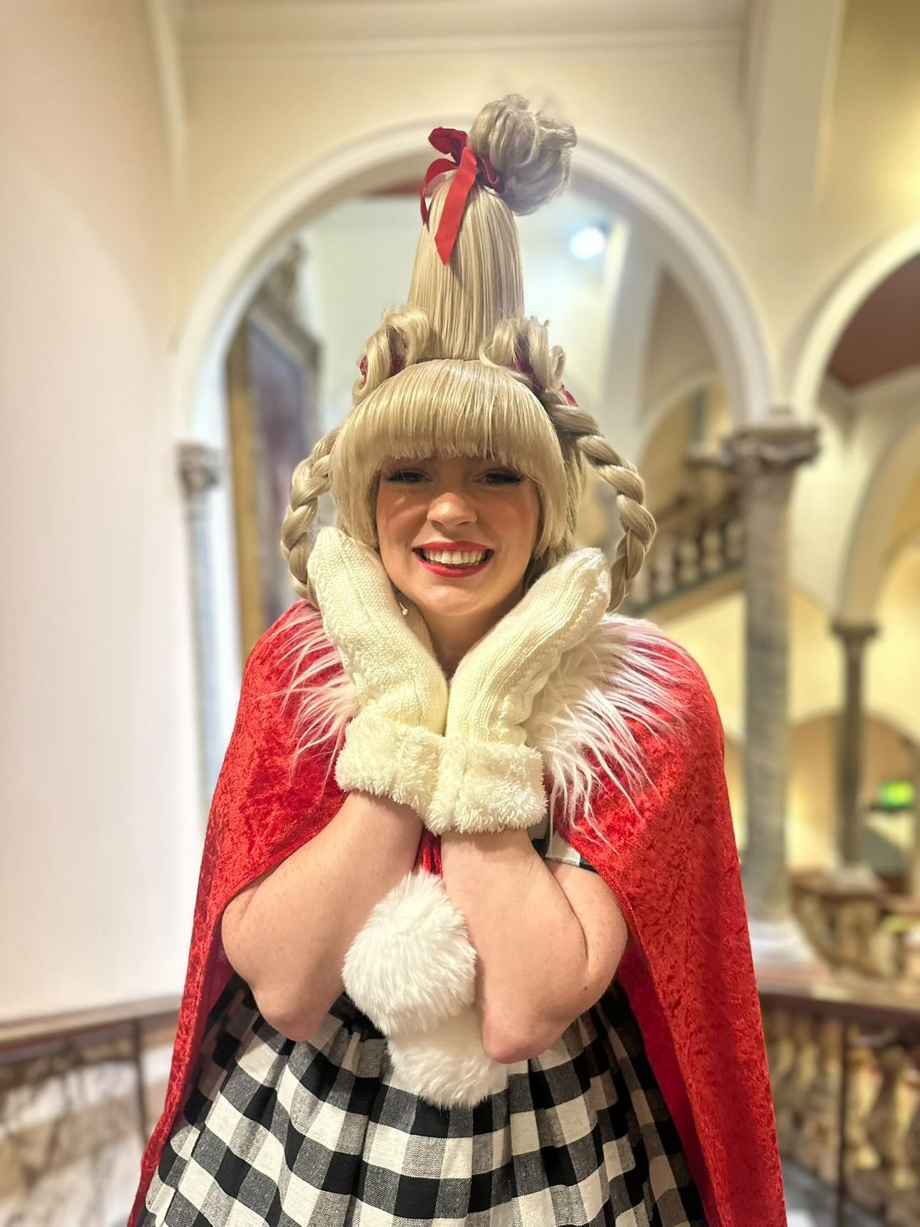 Woman with blonde hair styled with multiple curls and a large curl on top, wearing a red cape with white fur trim and large white gloves, standing inside a building with archways and columns.
