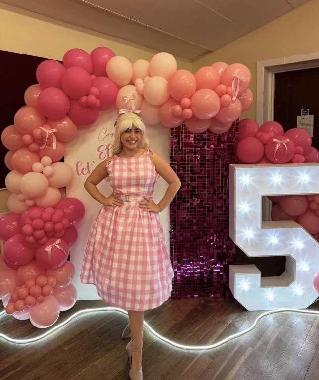 A woman smiling and posing in front of pink and white balloon decorations and a large illuminated letter 'C' at a celebration event