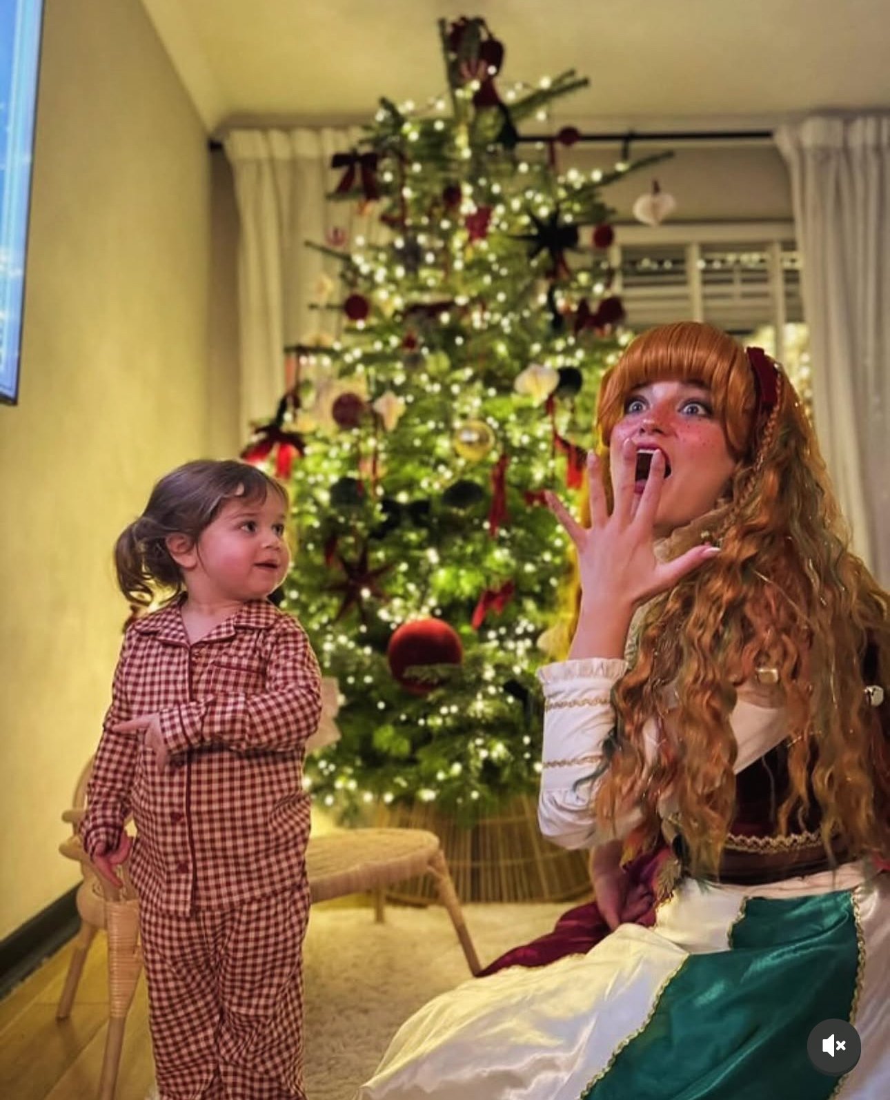 A young girl in red and white checkered pajamas standing near an adult woman with red hair, dressed in a fairy tale costume, in a living room decorated for Christmas with a decorated Christmas tree and curtains in the background.