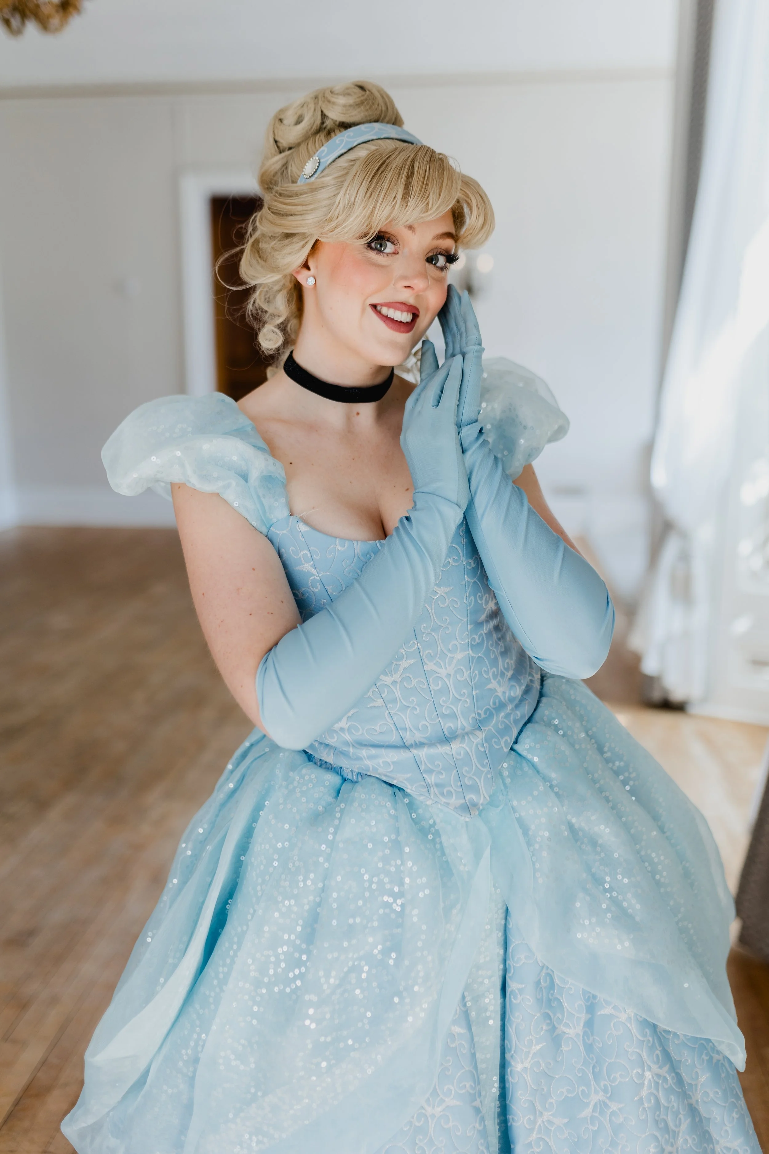 A woman dressed as Cinderella is smiling and posing with her hand near her face, wearing a blue ball gown, gloves, and a matching headband.
