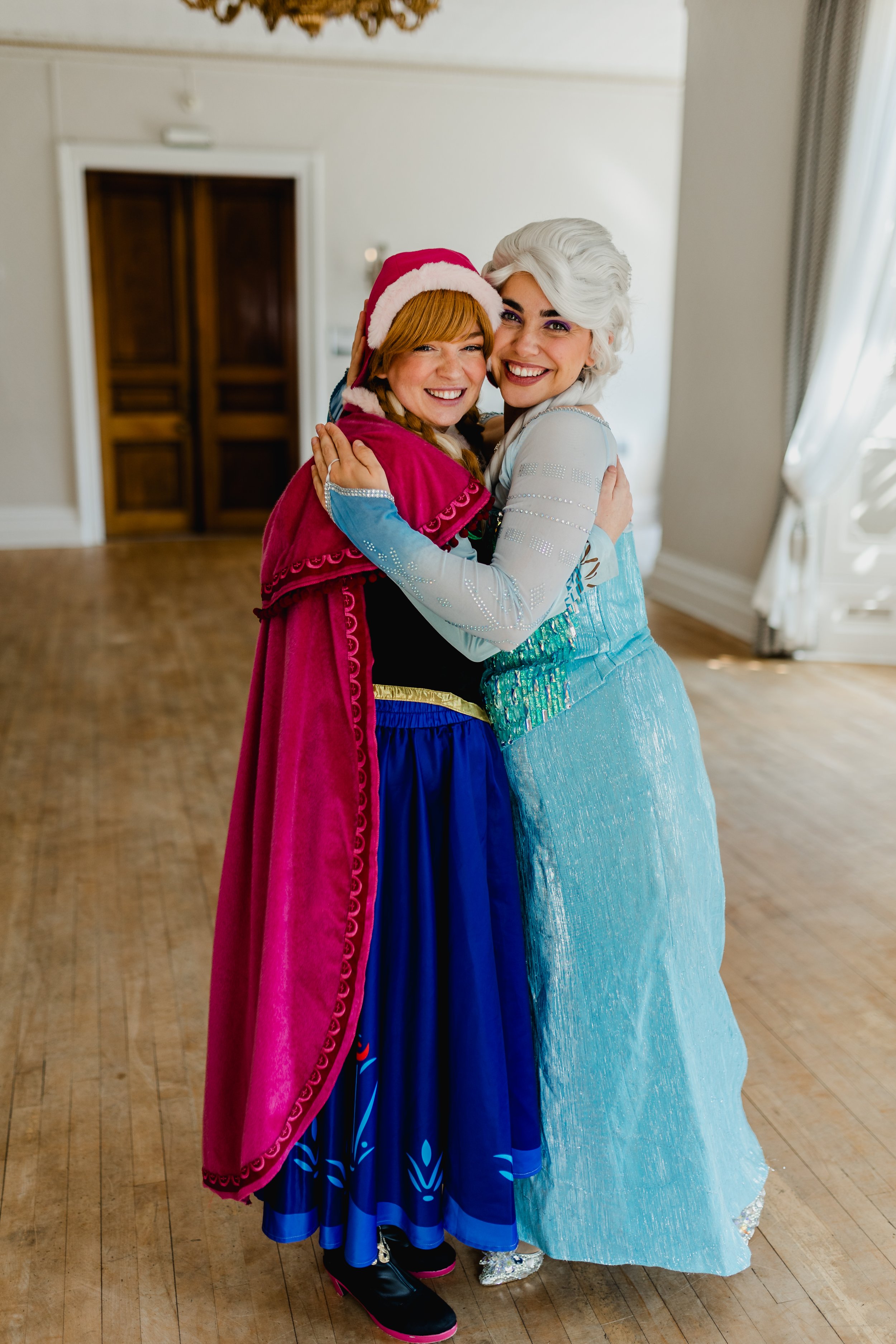 Two women in costumes hugging indoors, one dressed as Elsa from Frozen and the other as Anna, smiling. Princess Parties in Swanland