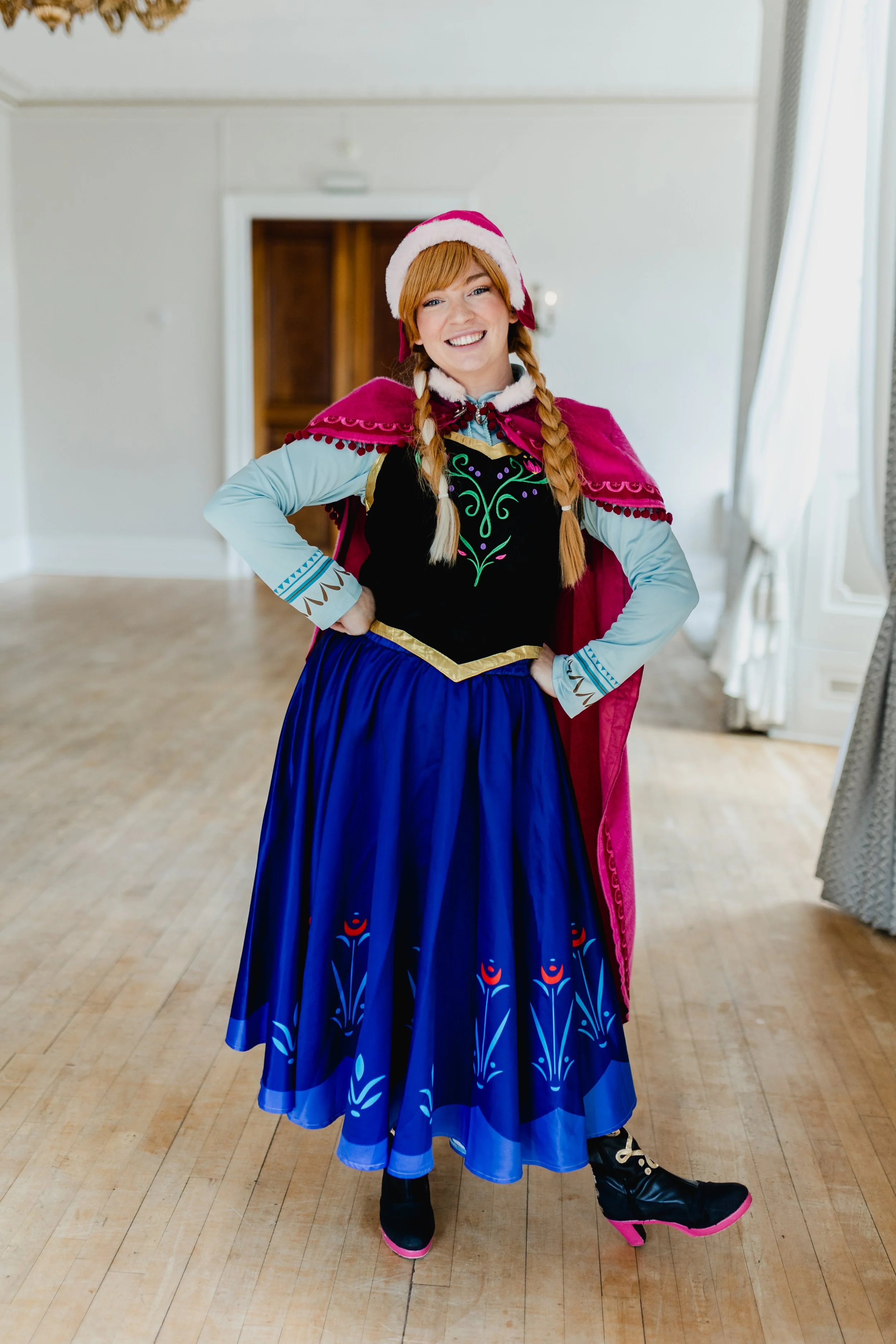 Woman in a cosplay of Anna from Frozen, wearing a pink and purple cape, blue skirt, and ice skates.