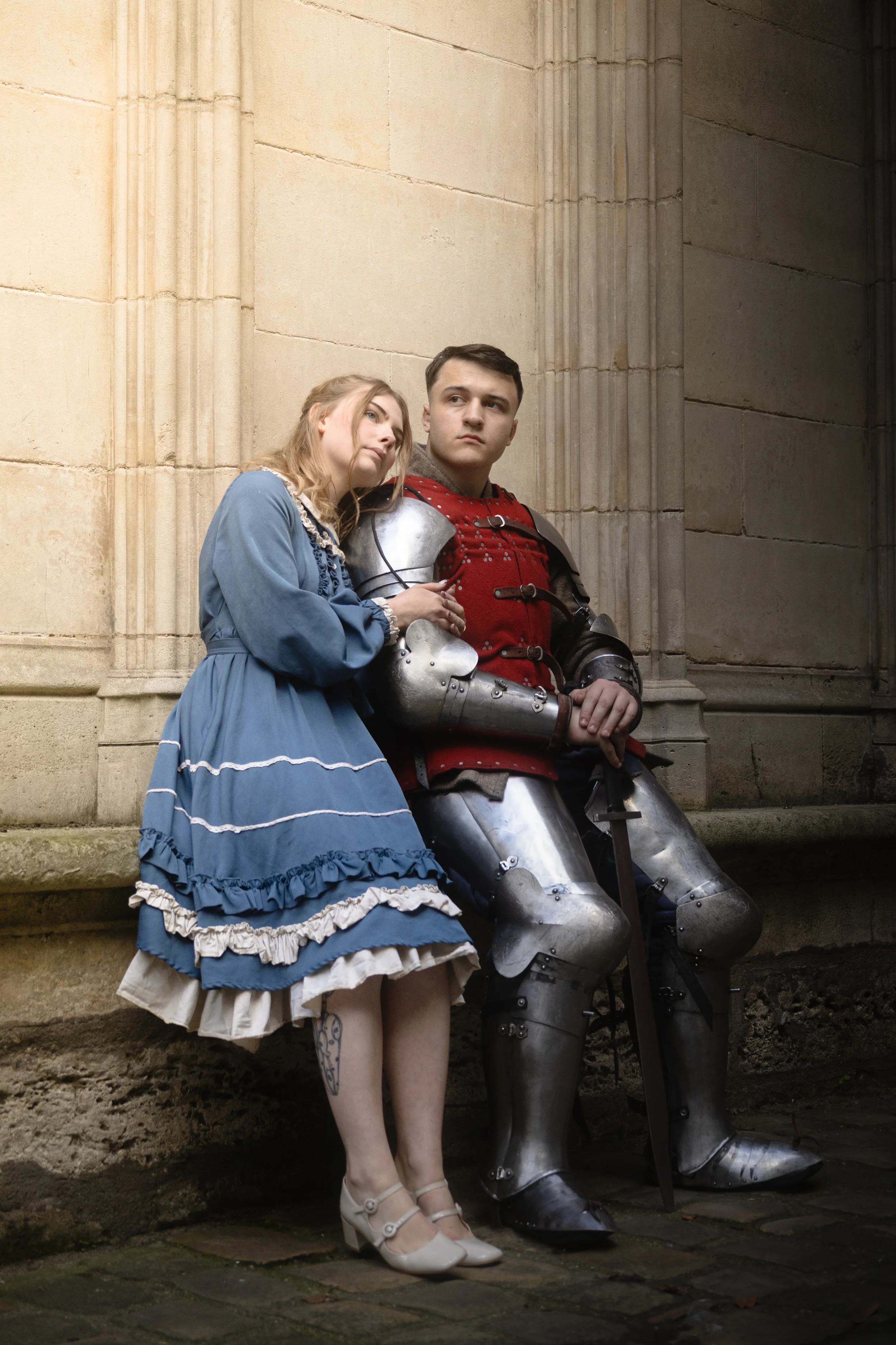 Un couple habillé en costumes médiévaux, un homme en armure métallisée et une femme en robe bleue, assis contre un mur en pierre d'une vieille bâtisse, dans une ambiance historique.