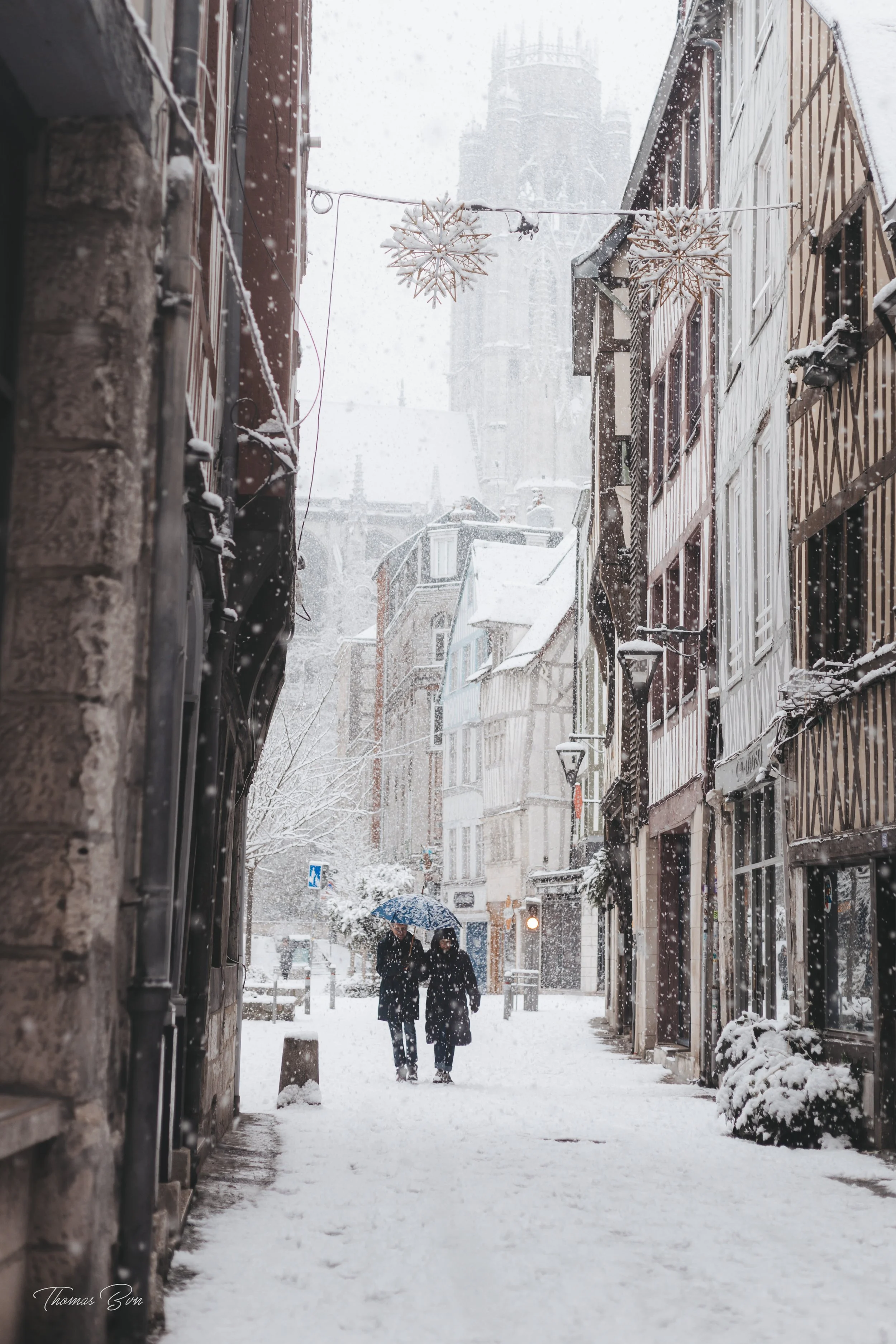 Rue pavée de neige avec deux personnes marchant sous un parapluie, bâtiments traditionnels et décorations de Noël suspendues. Neige abondante, forte en hiver.