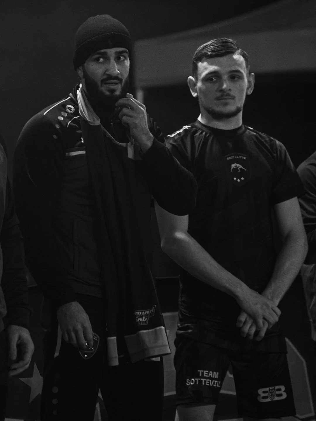 Deux jeunes hommes en tenue sportive dans une pièce sombre, l'un porte un bonnet et semble réfléchir tandis que l'autre regarde au loin.