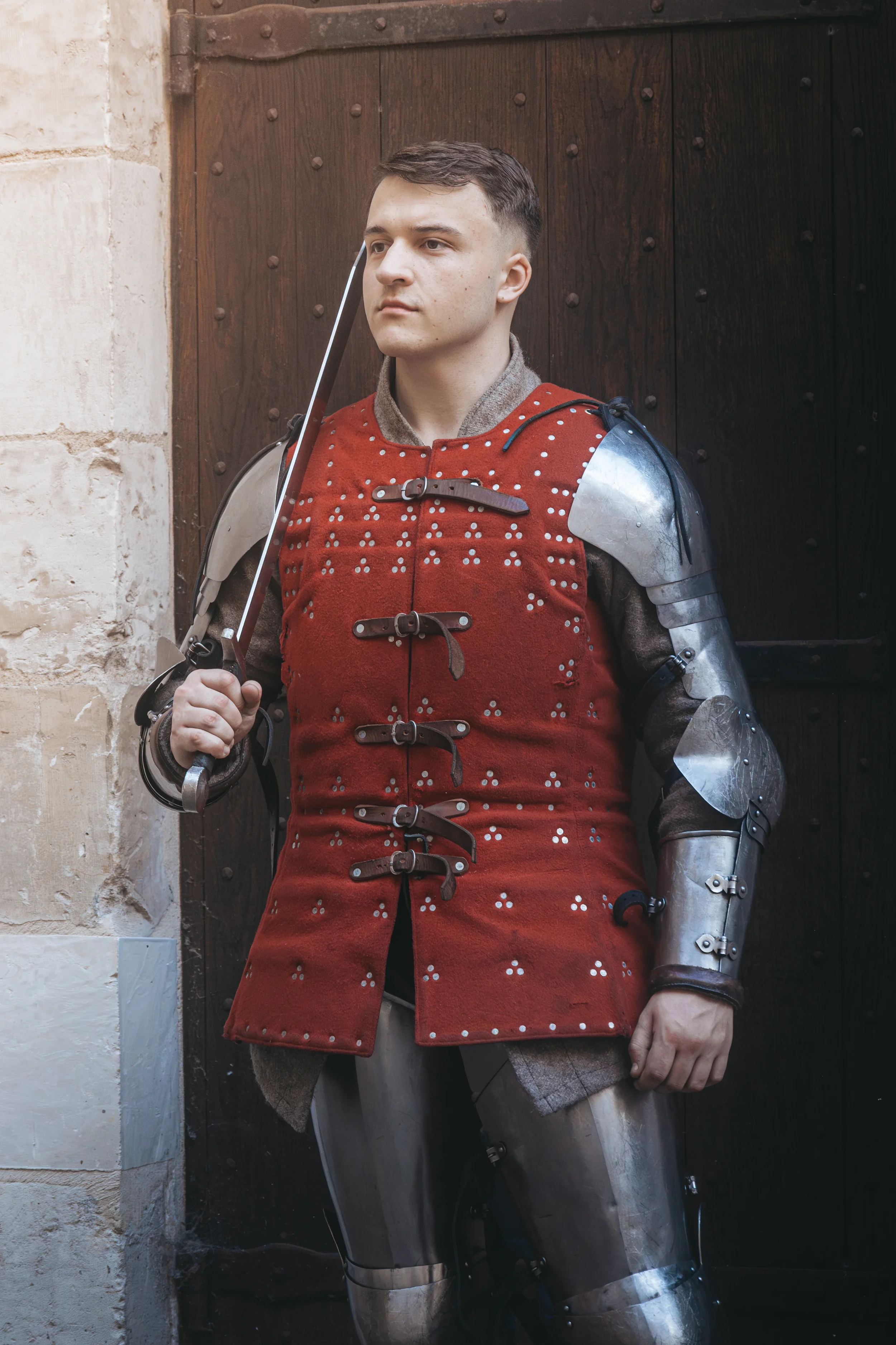 Jeune homme en armure médiévale avec une épée, debout devant une porte en bois, portant une cotte de mailles et une veste en tissu rouge avec des attaches en cuir.