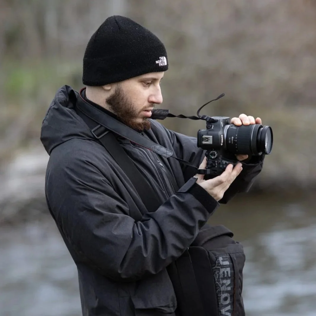 A man in black outdoor gear and a black North Face beanie looking at his camera outdoors near water.