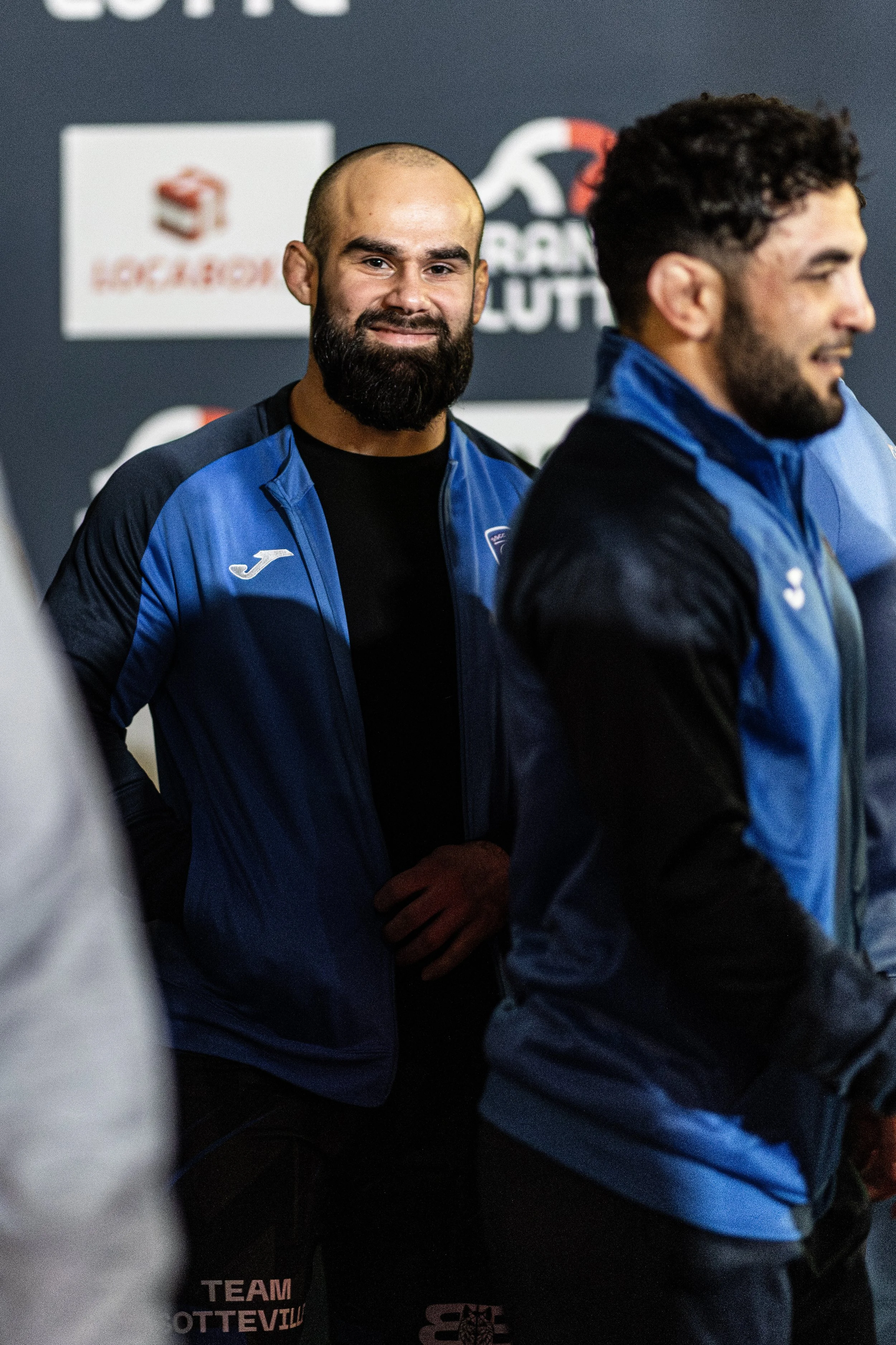 Deux hommes en vestes bleues avec des logos, assis ou debout dans un environnement sportif, avec un fond de logo et de texte.