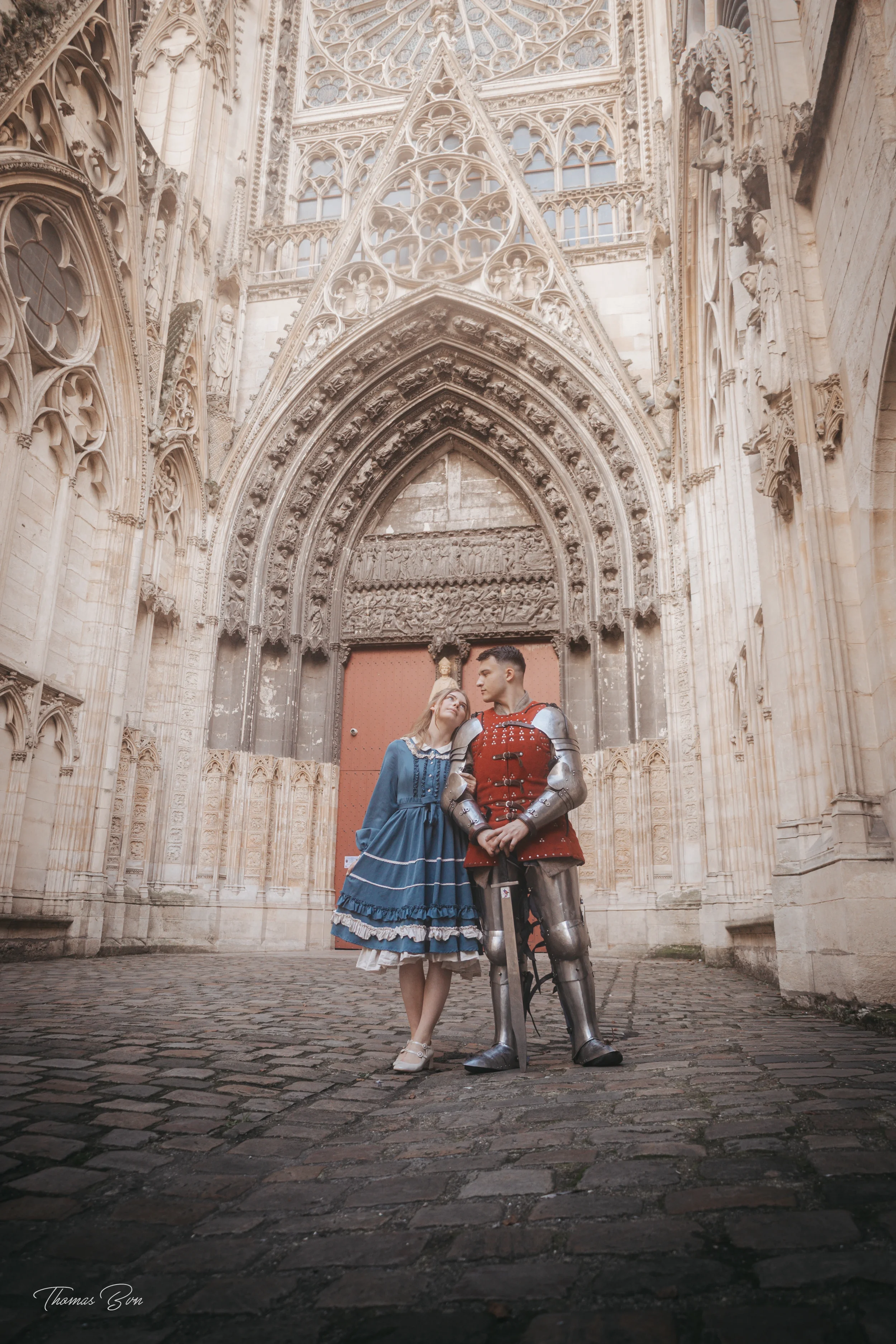 Une jeune femme en robe bleue et un homme en armure de chevalier posent devant la porte en pierre d'une cathédrale gothique