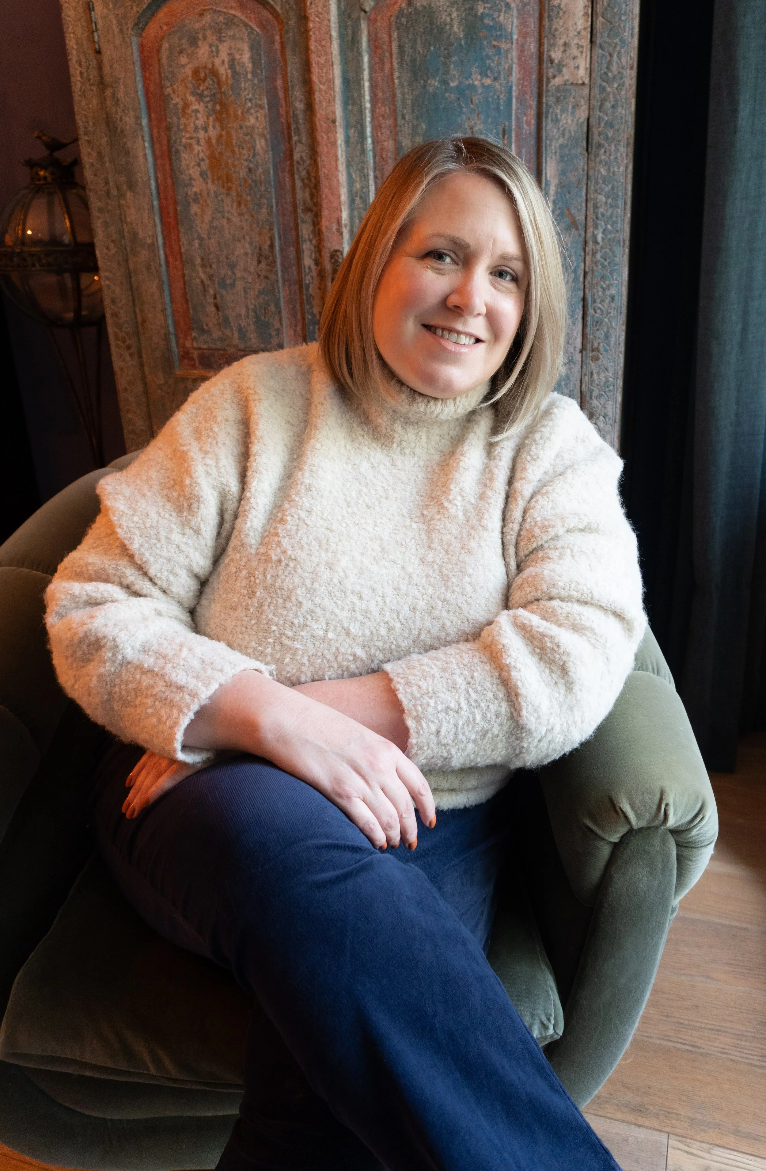 A woman with blonde hair wearing a cozy beige sweater, sitting on a dark green armchair, smiling at the camera in a room with old, textured wooden panels on the wall and a window with black curtains.