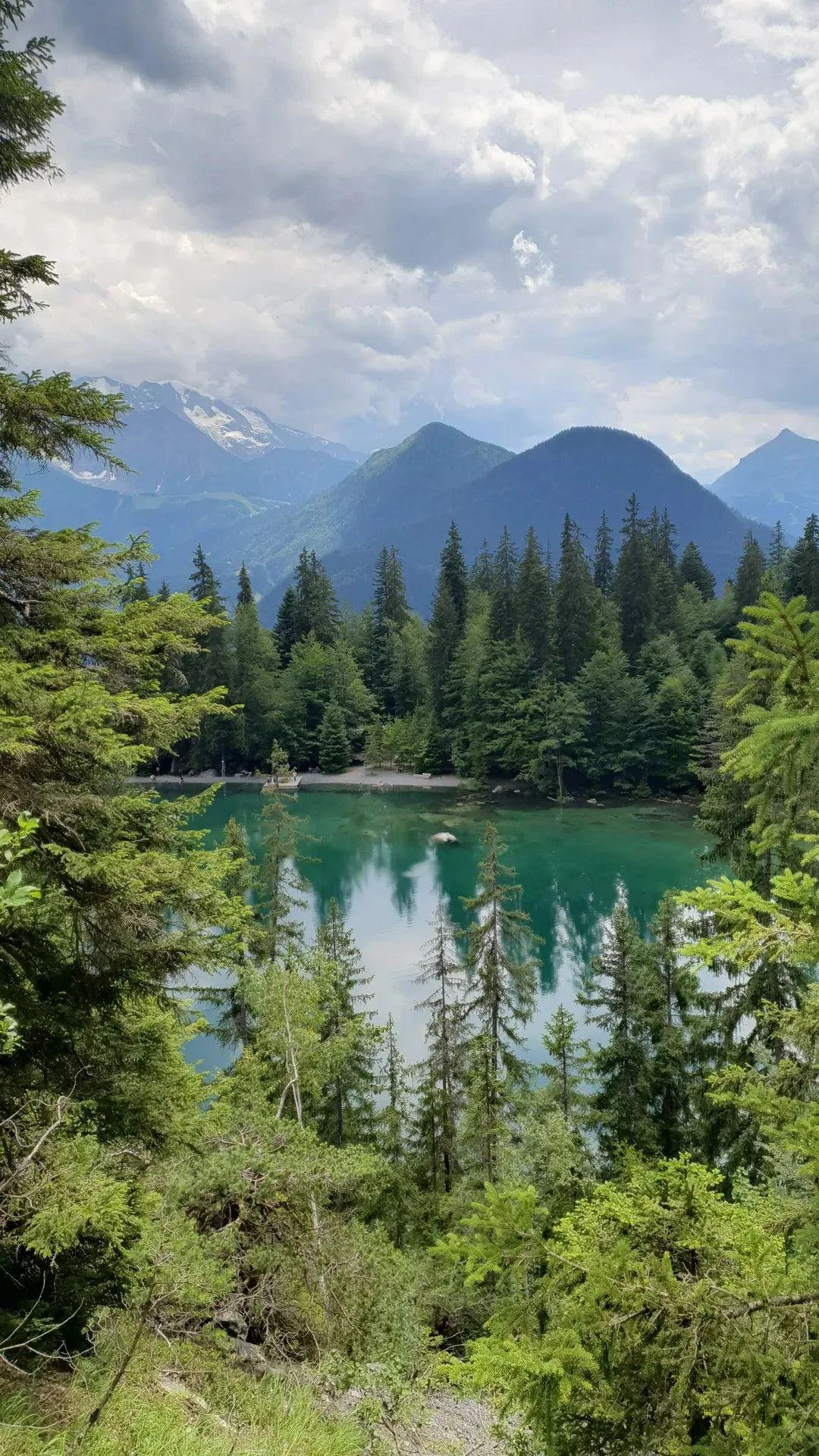 Lac vert alpin entouré de forêts de conifères avec montagnes en arrière-plan et ciel nuageux, Lac Vert, Passy, France