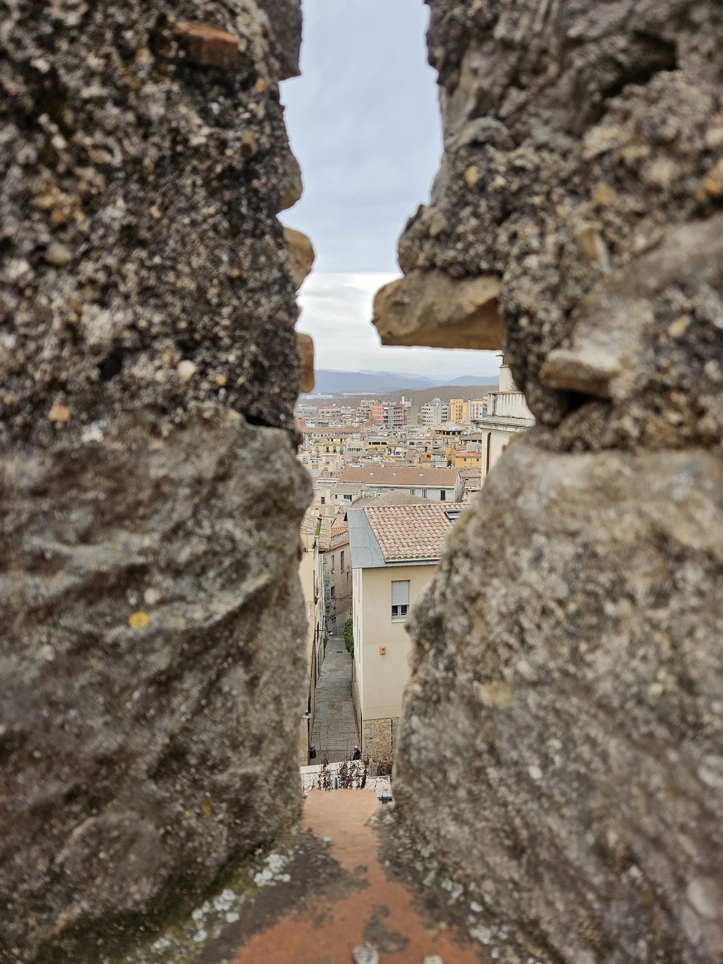 Vue d'une ville à travers une ouverture dans un mur en pierre, avec des bâtiments, des rues, et un ciel nuageux en arrière-plan, Remparts de Gérone, Espagne