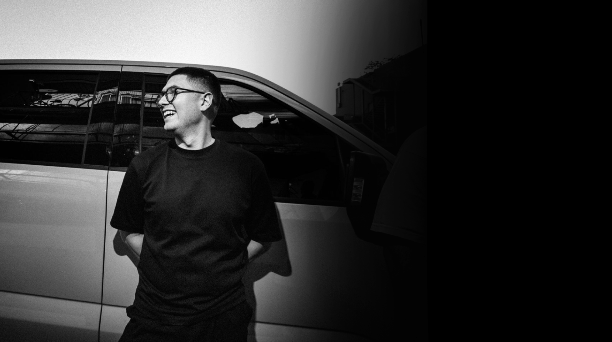A young man with glasses and a black shirt smiling and looking to the side, standing in front of a vehicle with a reflective window, during daytime.