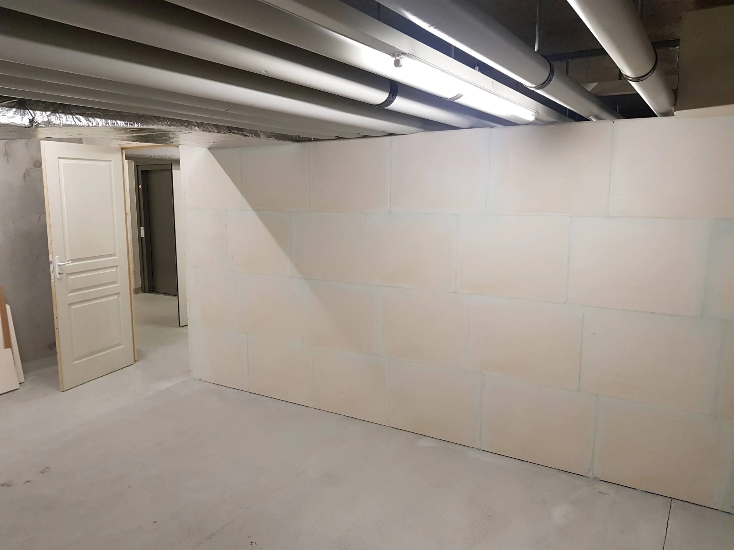 Une pièce en construction avec un mur en béton peint en blanc et un plafond perforé, des tuyaux visibles, une porte en bois blanc légèrement ouverte.