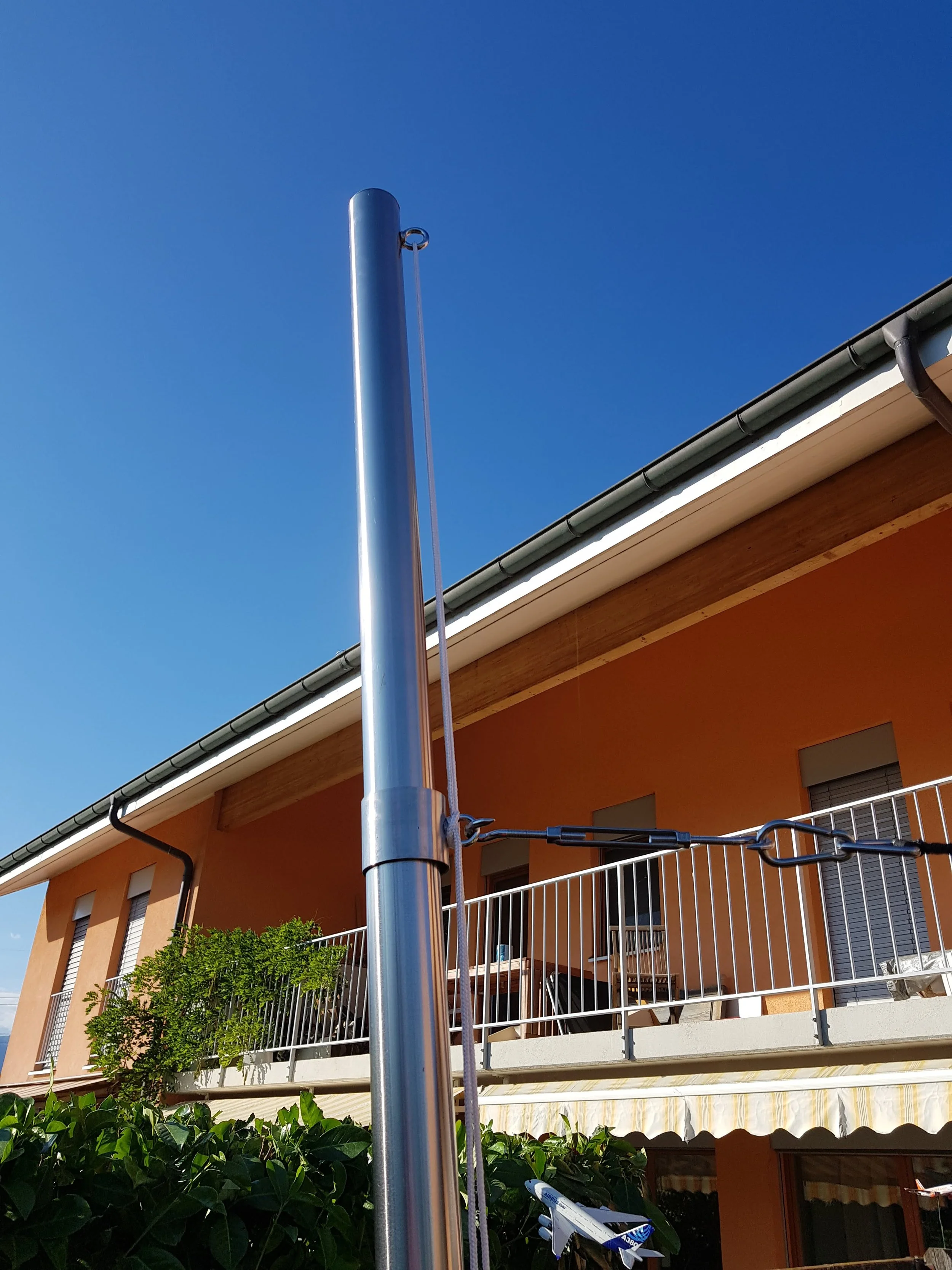 Un drapeau non hissé sur une hampe en acier, devant un bâtiment résidentiel avec un balcon orange, des volets fermés et un ciel bleu.