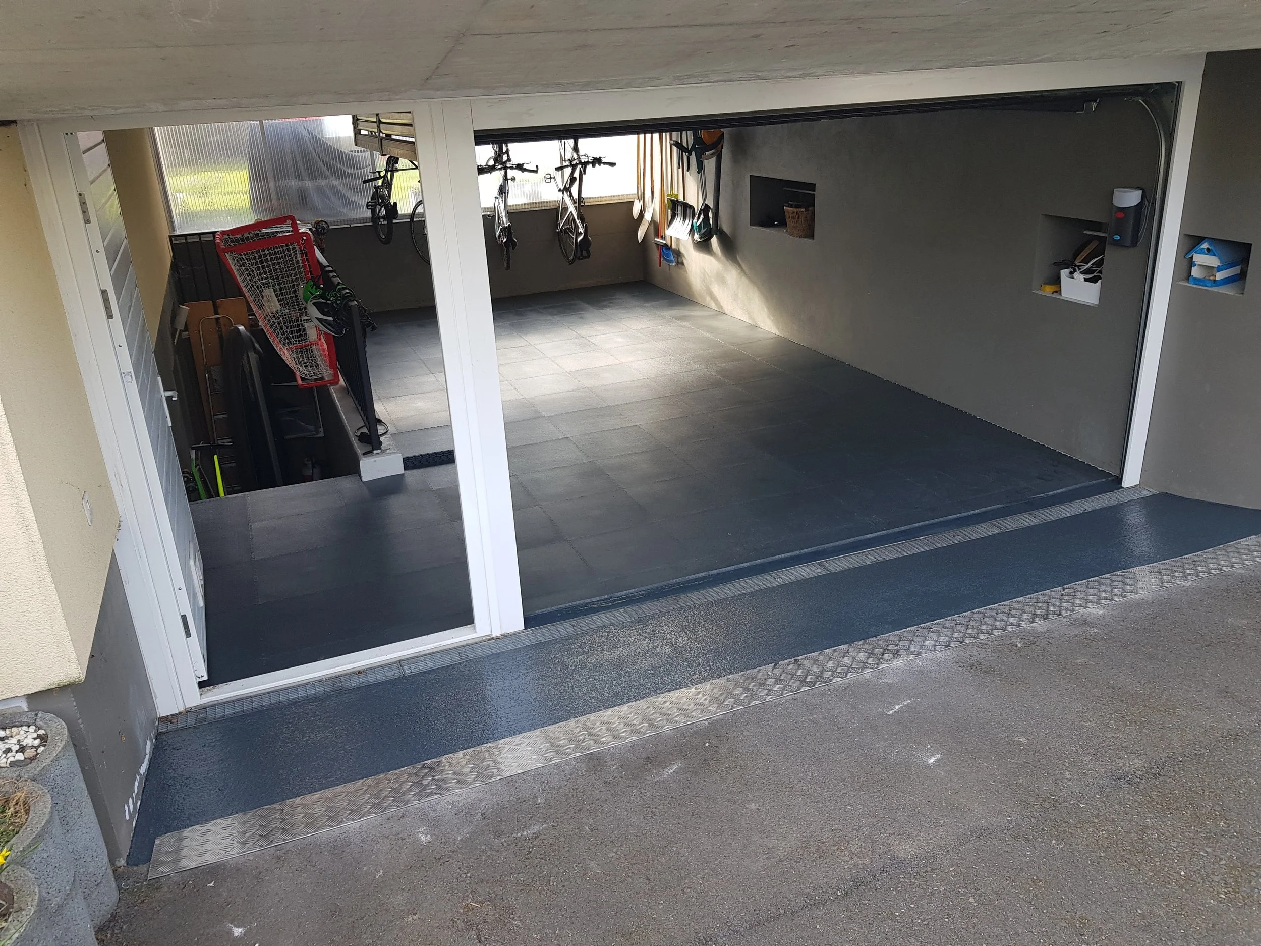 Garage souterrain avec deux vélos suspendus, une étagère avec outils, chaussures et autres objets, sol en carrelage gris, mur en béton peint, porte de garage enroulable partiellement ouverte, sol en asphalt à l'extérieur.