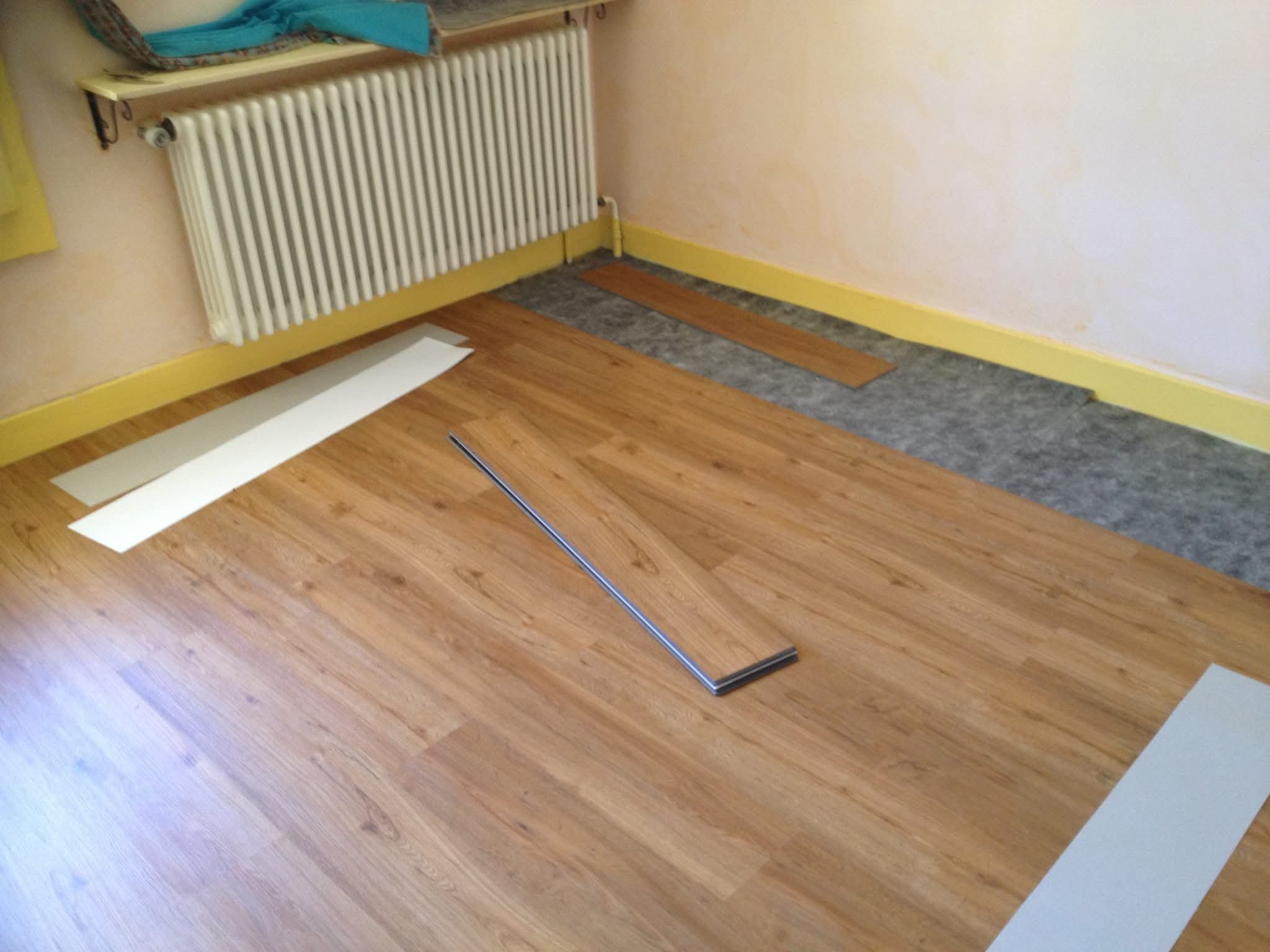 Une pièce en train d'être rénovée avec un plancher en bois partiellement installé, des bandes de protection, un radiateur, une étagère avec des objets dessus, et un mur beige avec un ruban de protection jaune.
