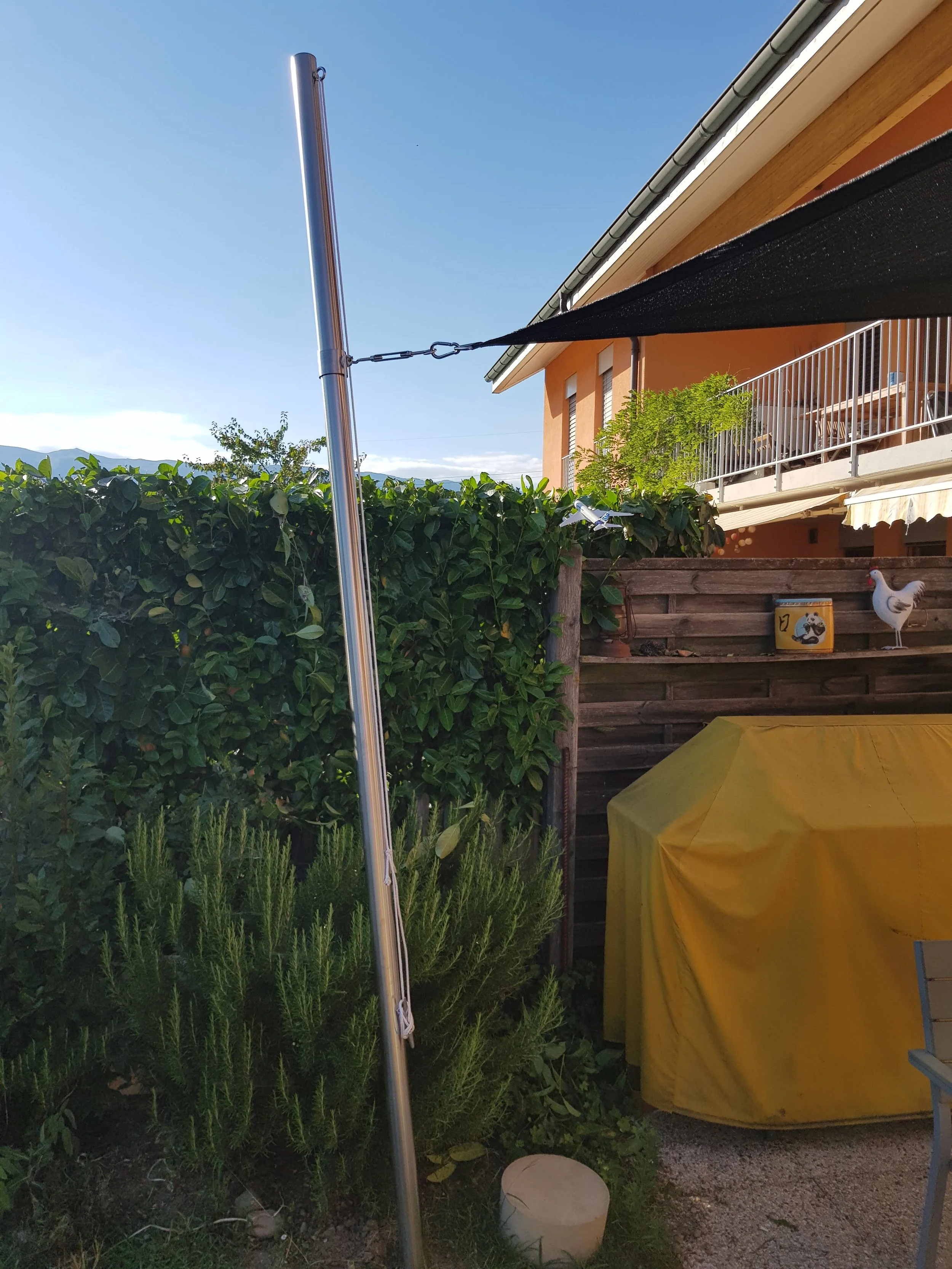 Balcon d'une maison avec un parasol noir, un objet jaune recouvert d'une housse, une clôture en bois, une plante verte, un panneau en forme de poule, et un mur orange avec des décorations