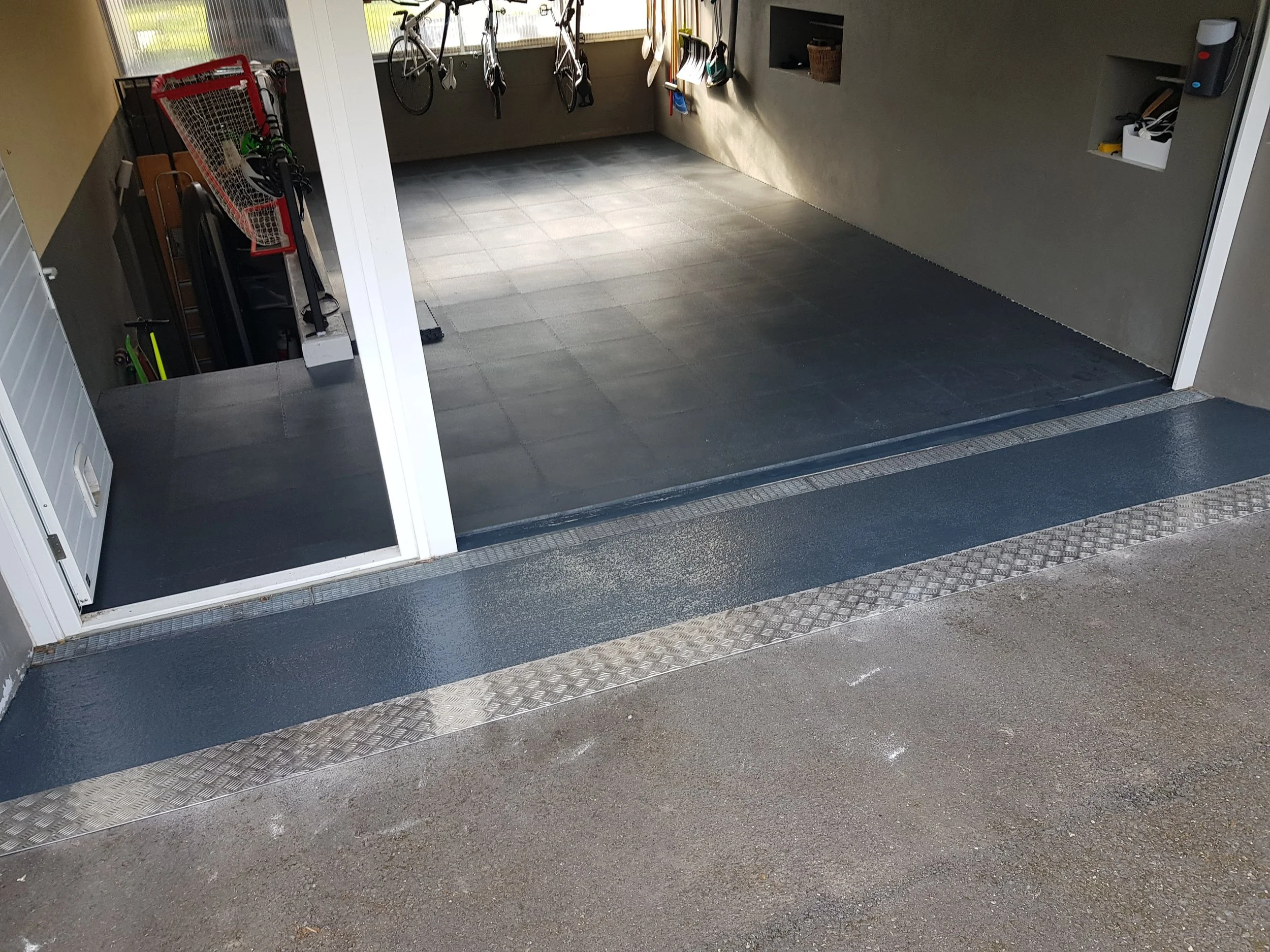 Saisie de garage avec sol en béton noir et équipements de vélo, outils de sport, et étagères. Plateforme accessible avec rampe en métal.