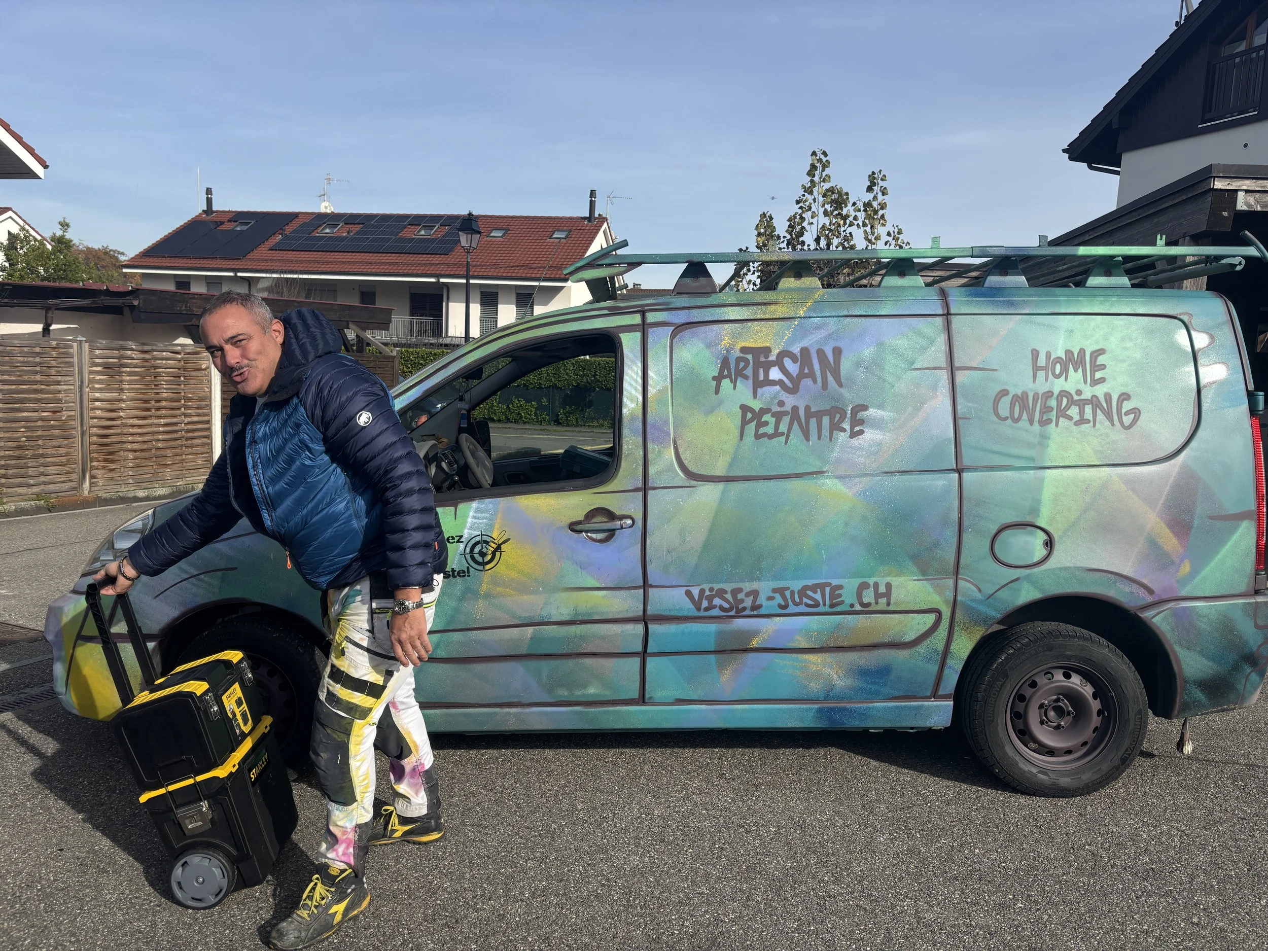 Un homme avec un sac à roulettes en plastique noir et jaune, debout à côté d'une petite voiture décorée avec un design artistique. La voiture affiche des textes en français sur ses côtés, comme 'ARTISAN PEINTRE' et 'HOME COVERING', ainsi que 'VISEZ-JUSTE.CH'. L'homme porte un pantalon coloré, une veste bleue, et des chaussures noires et jaunes.