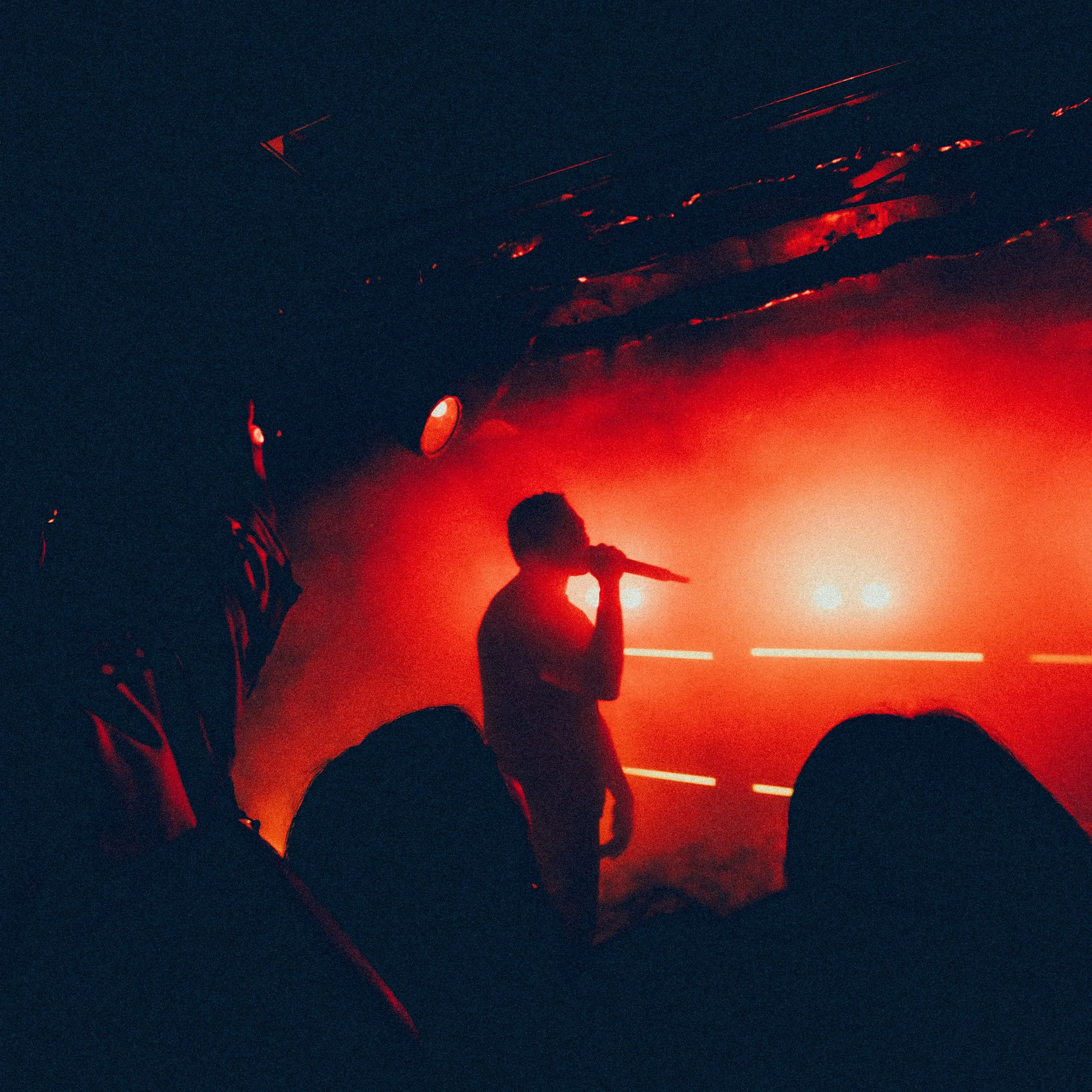 A person singing into a microphone on stage with red lighting and a smoky background, and an audience in the foreground.