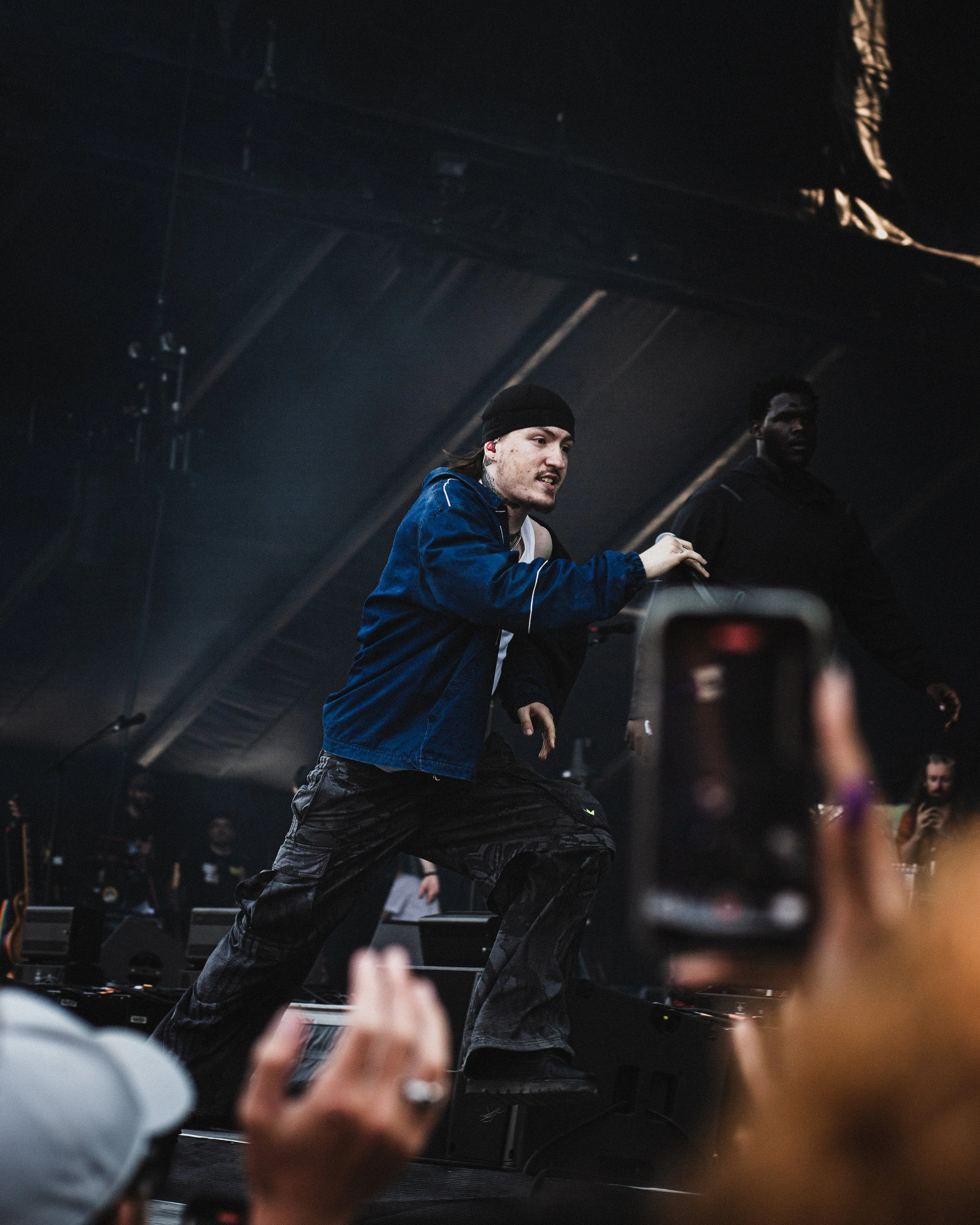 A performer on stage wearing a black beanie and blue jacket, dancing with a serious expression. Several audience members are visible, one taking a photo with a smartphone in front of the stage.