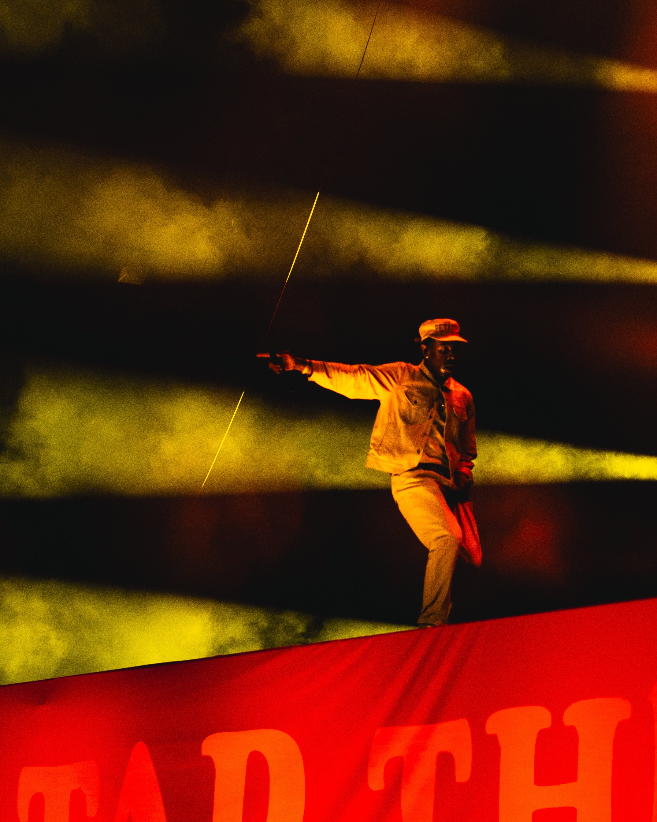 Man performing on stage with a microphone, wearing a cap, jacket, and pants, illuminated by dramatic lighting with fog effects.