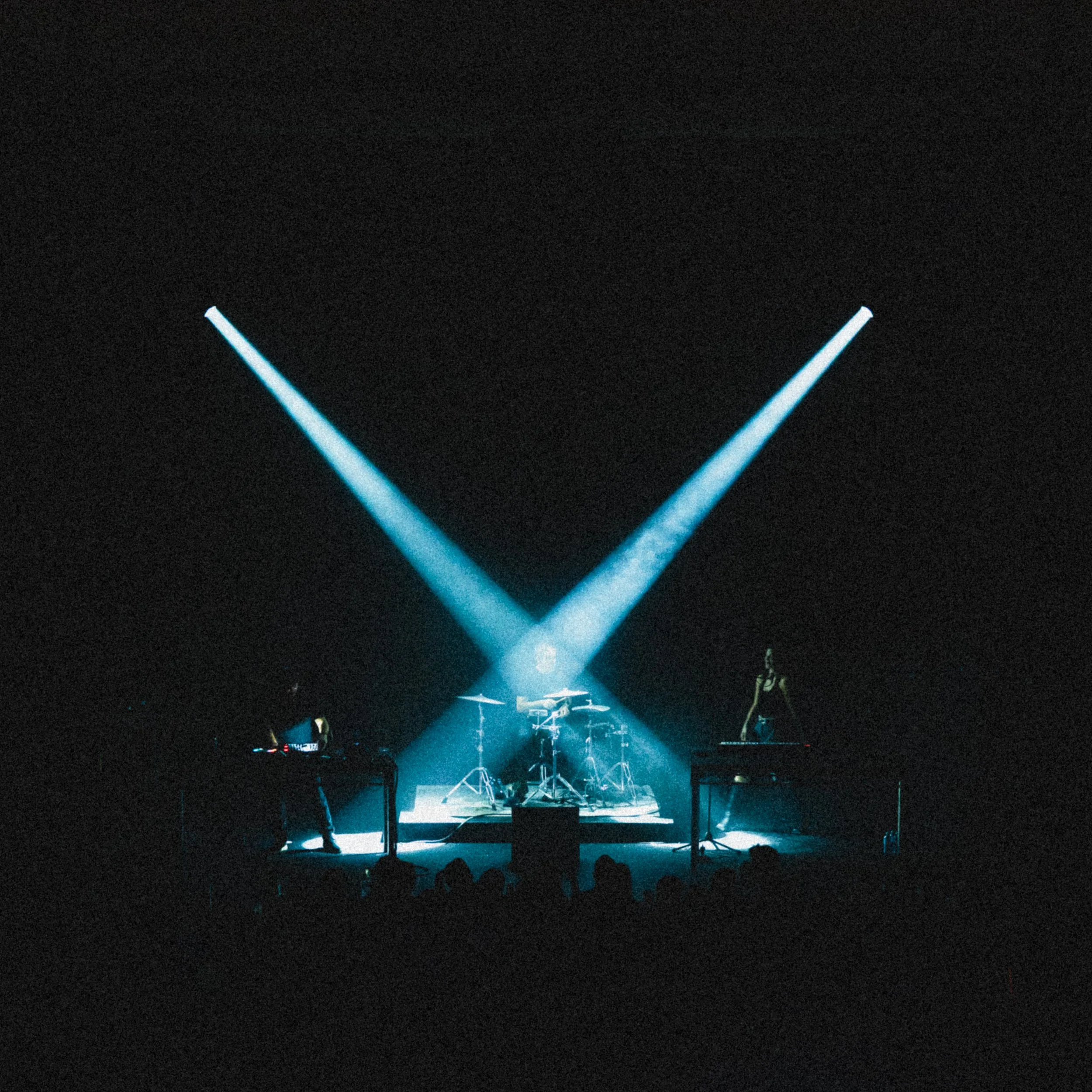 A dark stage with blue stage lights focused on musical instruments, including keyboards and a drum set, with silhouettes of audience members in front.