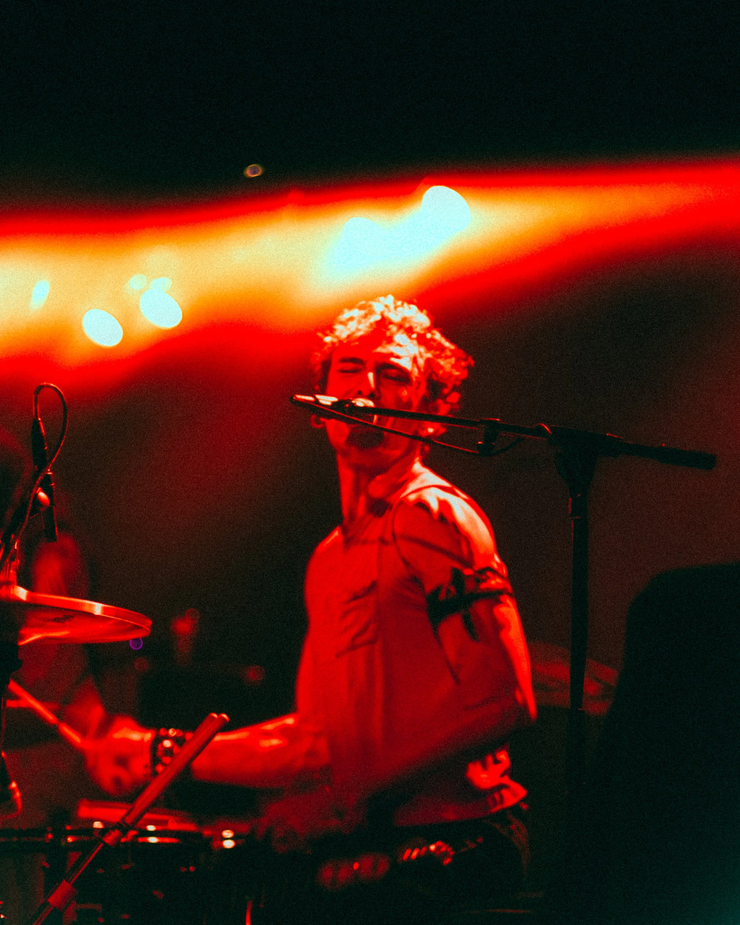 A musician with curly hair wearing a sleeveless shirt performing on stage, illuminated by red stage lights.