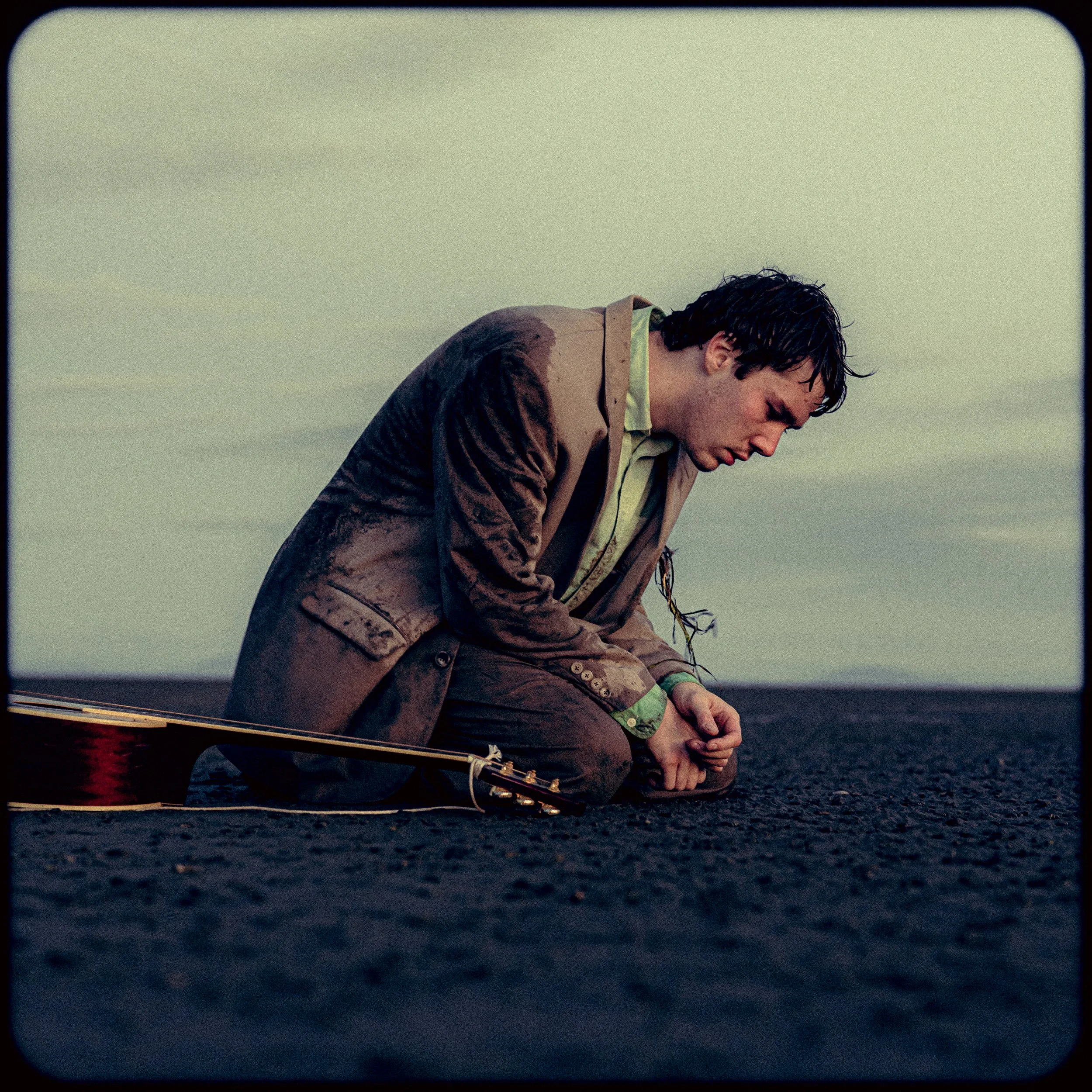 A man kneeling on dark ground, looking down with a somber expression, with a guitar lying beside him.