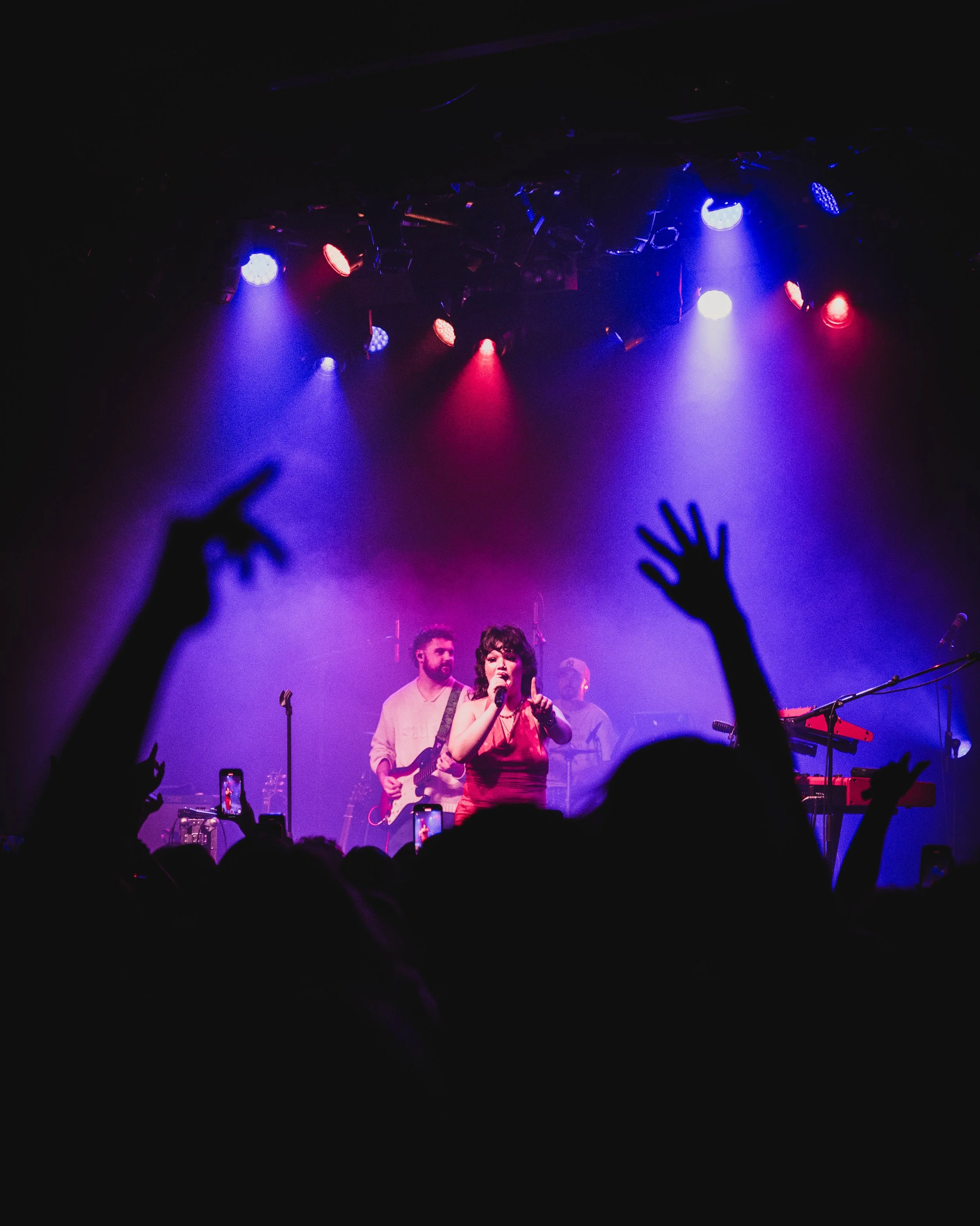 A live music concert with a singer in a pink dress performing on stage with band members, under colorful stage lights, with audience members raising hands and recording the performance with their phones.