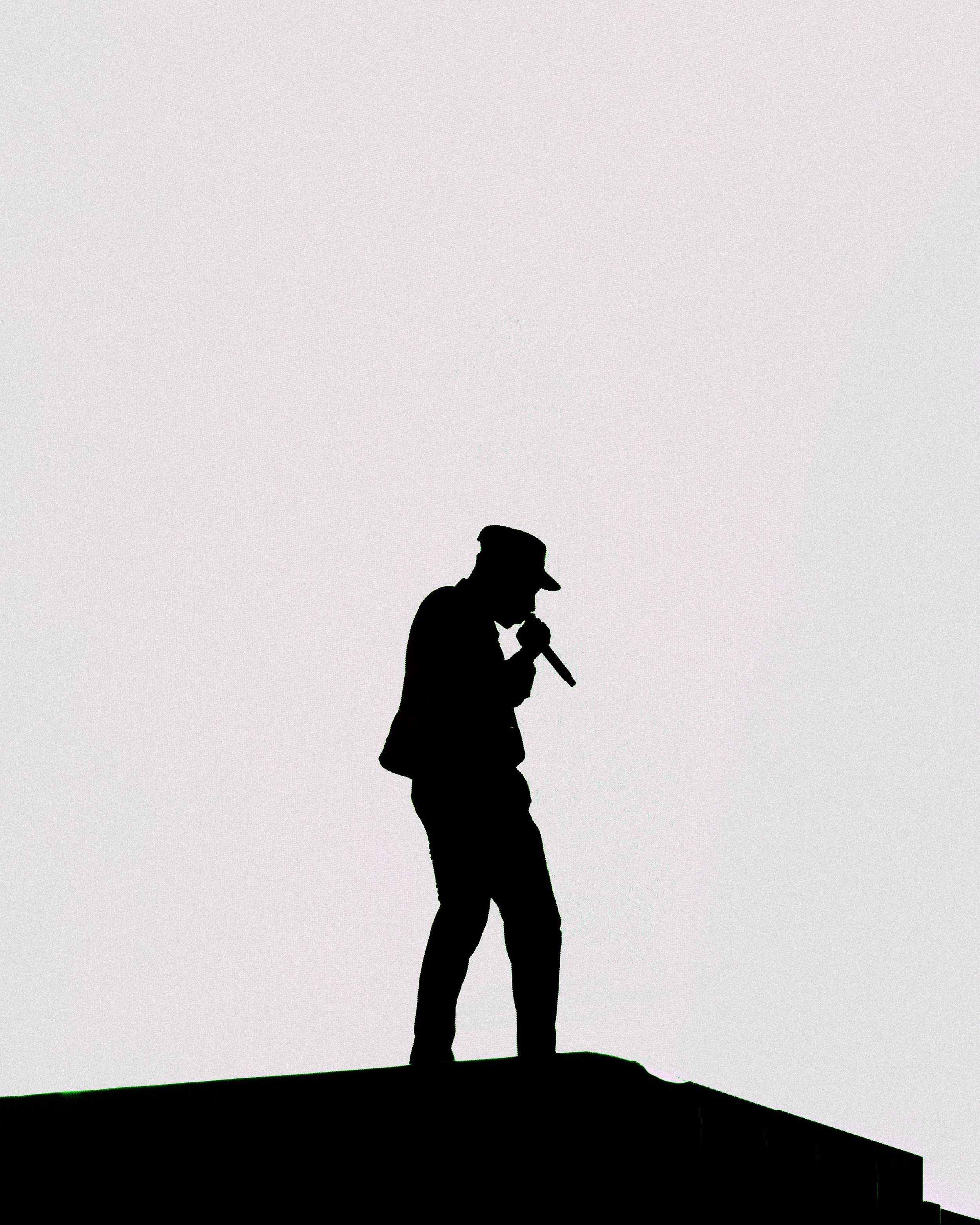 Silhouette of a man holding a microphone and a piece of paper, standing on a rooftop against a plain white sky.