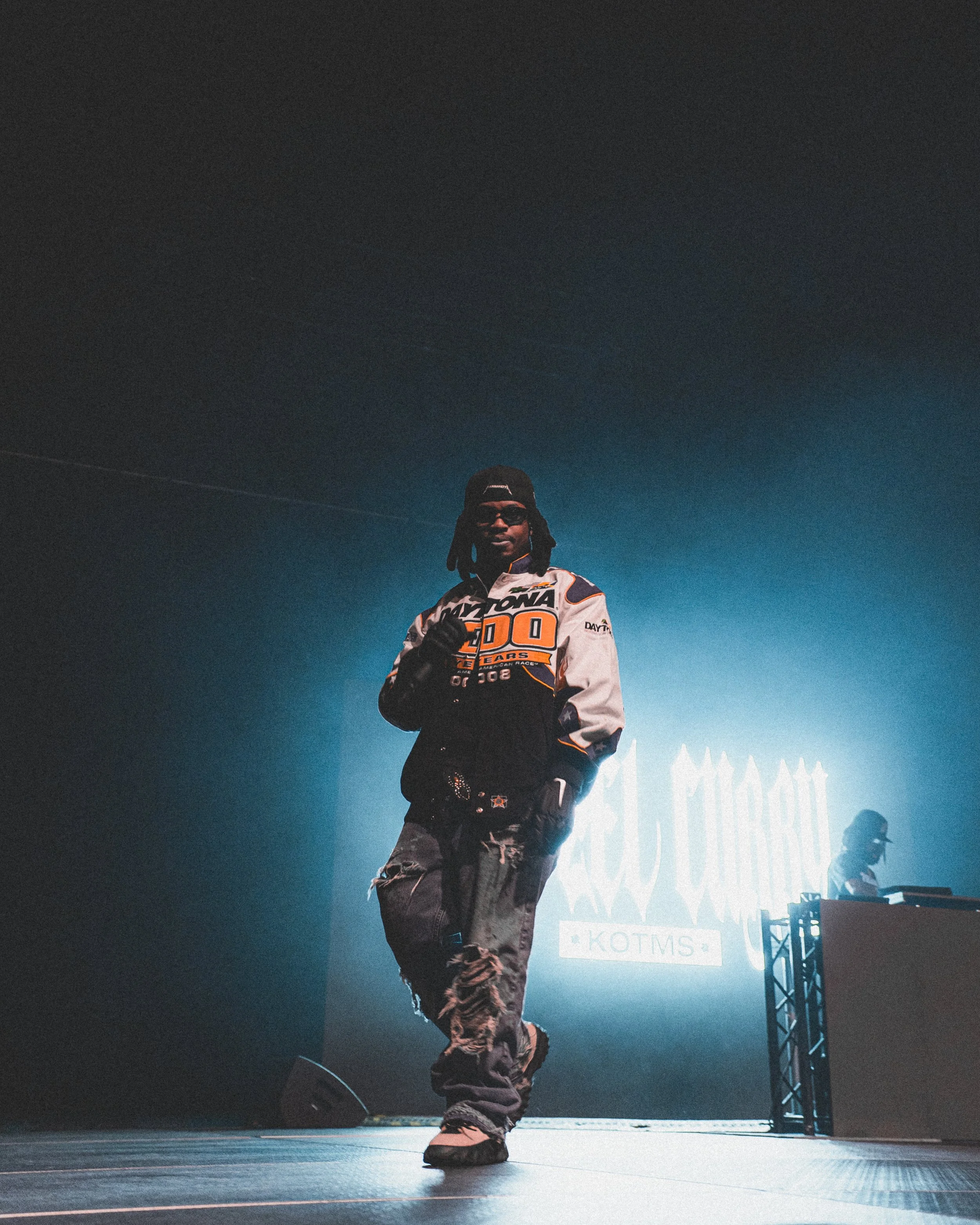 A person standing on stage at an event with a large bright logo in the background, wearing sunglasses, a black and white racing jacket, ripped jeans, and sneakers, with a DJ or musician in the background.